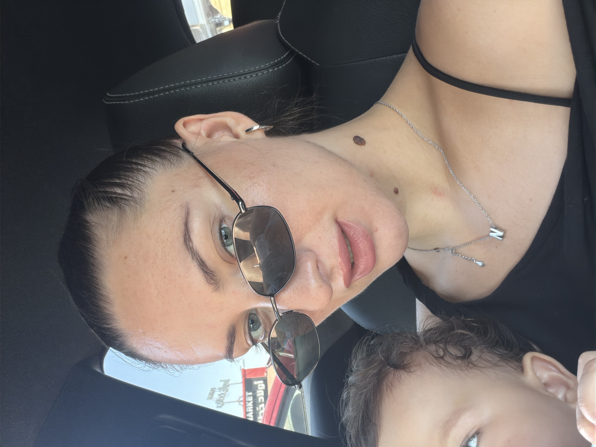 This is a medium shot, close-up, selfie-style photo taken inside a car. The main subject is a woman with light skin, green eyes, and dark hair pulled back. She is wearing sunglasses, a black tank top, and a silver necklace with a pendant shaped like the letter 'N'. A mole is visible on her neck. Next to her, a baby with curly brown hair and light skin is partially visible, looking to the left. In the background, outside the car, a red vehicle and a sign that reads "MARKET" in English and Arabic can be seen. The lighting suggests it is daytime. This scene is consistent with an outdoor setting in Sharm El Sheikh, Egypt, given the style of the signage and the generally sunny weather typical of the region.