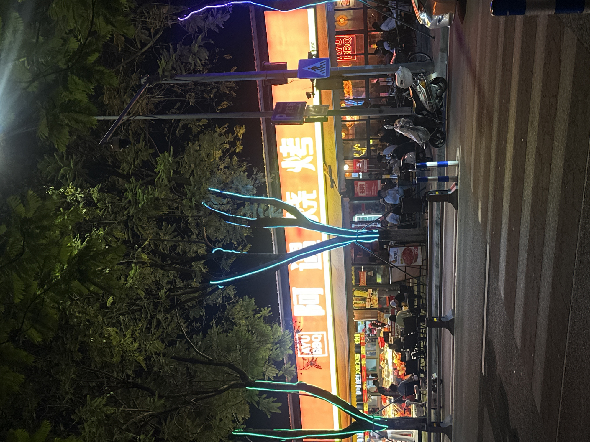 The image depicts an outdoor scene at night in the city of Chongqing High-tech Zone, China. A brightly lit restaurant with an orange awning is visible, featuring the text "AYU BBQ" and Chinese characters. Neon blue lights are wrapped around the trees in front of the restaurant, illuminating the area. Several patrons are seated at tables outside the restaurant, some appearing to be eating or conversing. A crosswalk is present in the foreground, and two scooters are parked on the sidewalk. Streetlights and the glow from the restaurant cast light on the scene. The overall atmosphere suggests a lively evening at a local eatery.