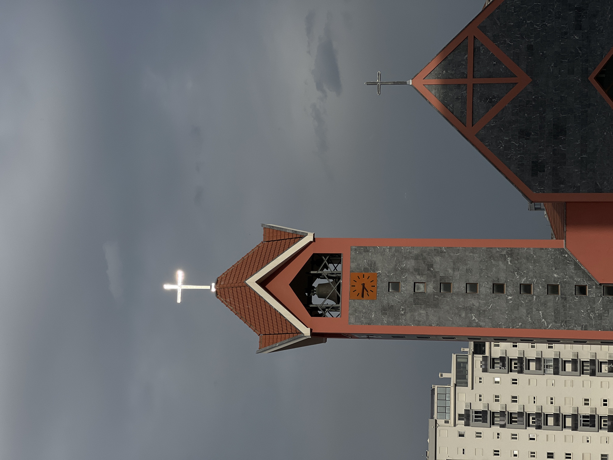 A low-angle, rotated view shows the Catholic Church of St. Peter the Apostle in Bar, Montenegro, under a cloudy sky. The church features red and grey marble elements. The bell tower has a clock face and a cross on top. A modern residential building is visible behind the church.