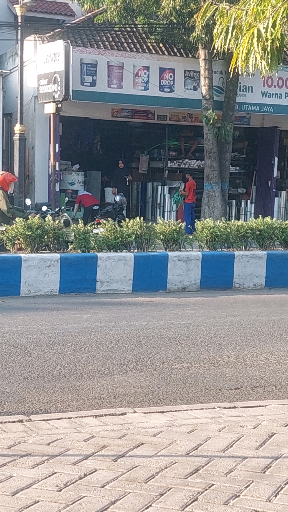 The image depicts a street scene in front of a shop in Nologaten, Indonesia. The shop has a sign above its entrance with the text "lemkote" and advertisements for paint products, including cans labeled "NO DROP". Inside the shop, various items are displayed, including what appear to be pipes and building materials.
In the foreground, a street with a paved sidewalk and a concrete curb painted with alternating blue and white blocks is visible. A person wearing a red shirt and blue pants is standing near the shop entrance, holding a green hose. Another person in a red shirt is bending down next to a motorcycle. A person wearing a dark hijab and a black cloak is standing further inside the shop entrance. To the left, a motorcycle with a red helmet on its seat is parked. Trees with lush green foliage are visible above the shop. The overall scene appears to be an outdoor commercial area in a populated locality.