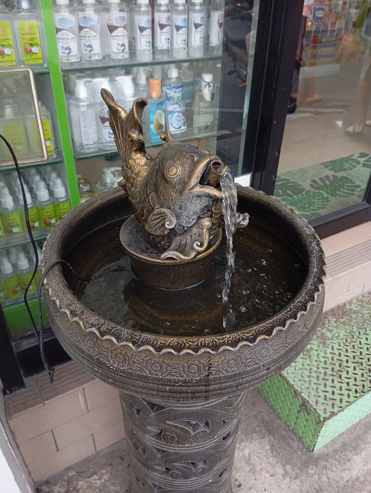 A close-up, eye-level shot captures a decorative fish fountain situated on a pedestal outside a shop in Karon, Thailand. The fountain's basin, a weathered bronze-colored circular bowl with ornate floral patterns around its rim, is filled with dark water from which a stream cascades from the open mouth of a bronze-colored fish ornament. The fish, positioned centrally atop a smaller bowl, is intricately detailed, with scales and fins clearly visible. A black electrical cord for the fountain pump is visible trailing from the water. Behind the fountain, a glass display case reveals shelves stocked with various bottles, many appearing to be coconut oil products, evidenced by labels featuring coconuts. A bright green metal platform with a diamond plate pattern is positioned to the right of the fountain and pedestal. The ground beneath is a mix of concrete and grey paving stones. In the background, through the shop window, blurred figures of people can be seen, indicating a public area.