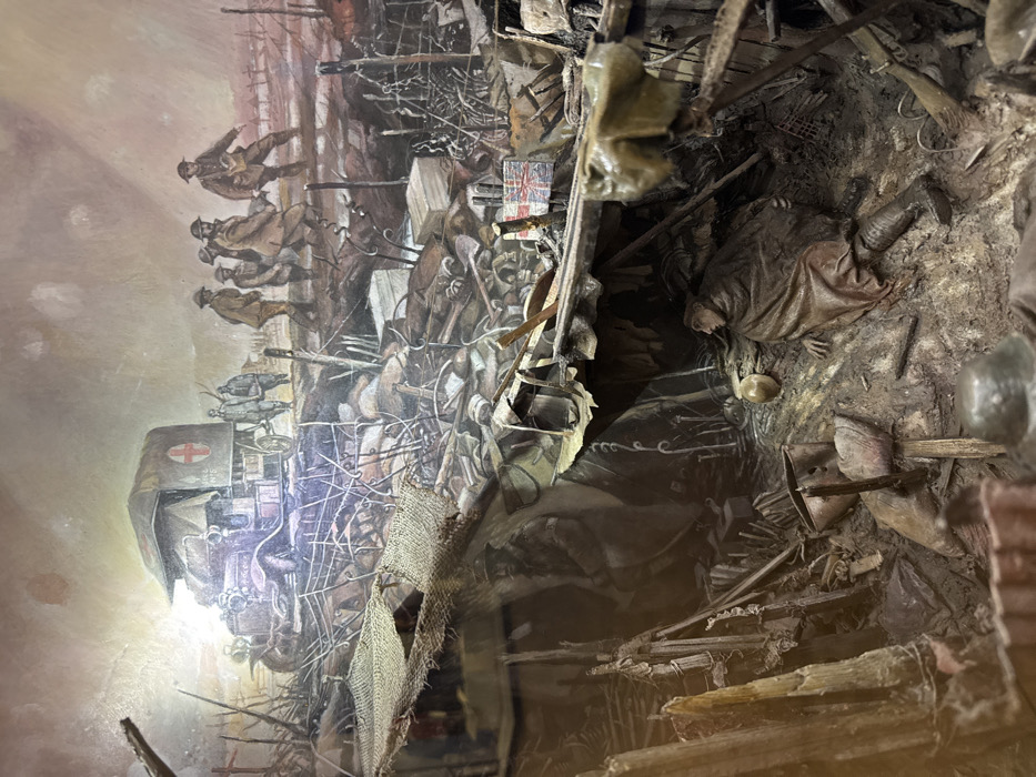 The image is a diorama depicting a scene from World War I. It is located in the Imperial War Museums, located in London, United Kingdom at latitude 51.497 and longitude -0.176. The diorama shows a group of soldiers in the foreground, in a trench, with a background painting of more soldiers behind a barbed wire fence. The scene features an ambulance, a stretcher, and a dead soldier, with a Union Jack flag in the background. The soldiers in the painting are carrying a stretcher toward the ambulance, while the diorama shows a dead soldier lying on his back in the mud, his arms outstretched, surrounded by debris and wreckage. The scene is set in a muddy trench, with barbed wire and other debris surrounding the figures. The diorama uses various materials, including plaster, wood, and metal, to create a realistic portrayal of the scene. The lighting is dim and the overall tone of the image is one of darkness and despair.