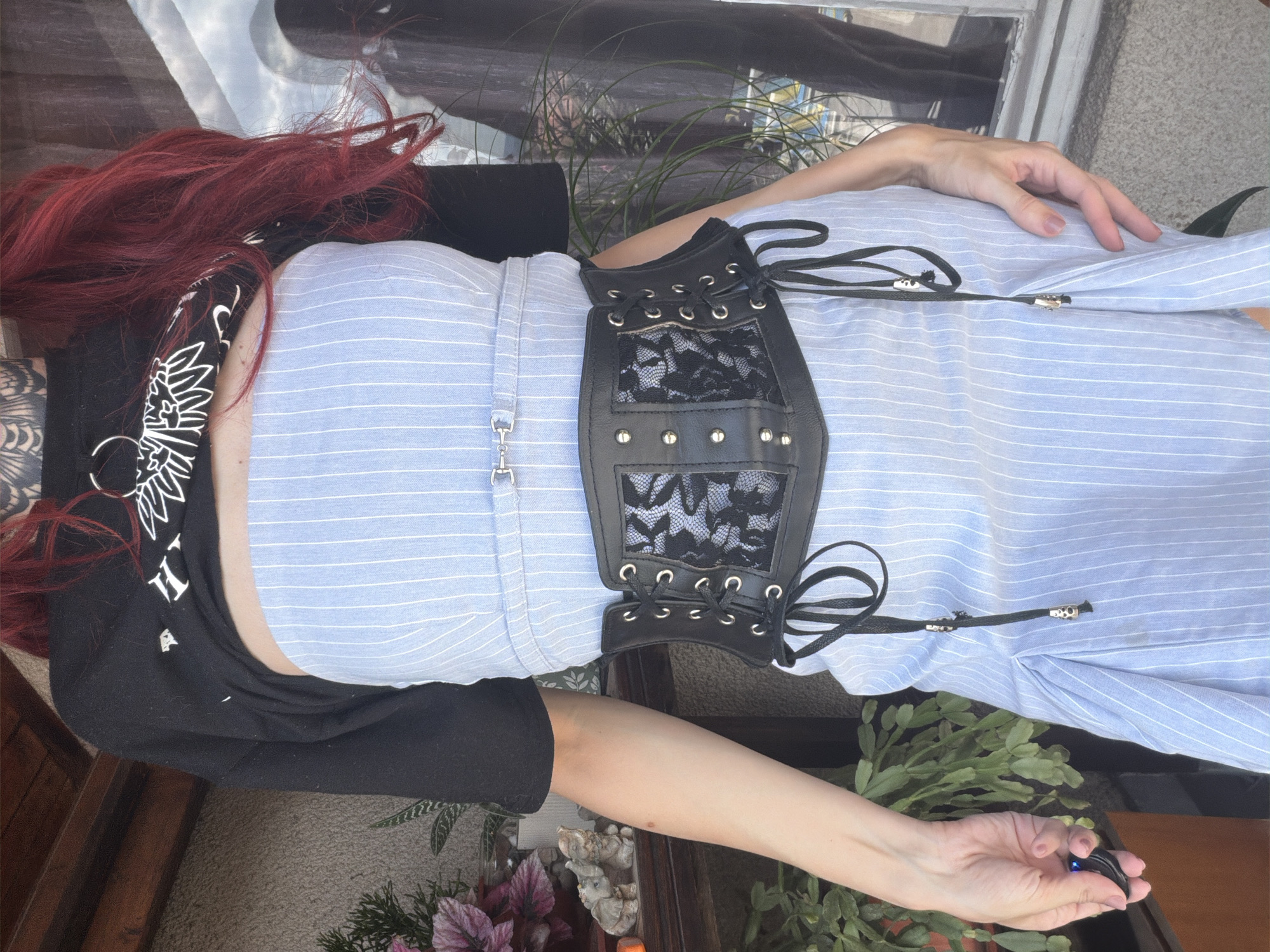 A person with red hair and tattoos on their upper body is standing in front of a house with a tiled facade, possibly in Svilengrad, Bulgaria. They are wearing a black cropped sweatshirt over a light blue pinstriped strapless dress. A black corset belt with lacing and black lace detailing is cinched around their waist. The person is holding a small black electronic device in their left hand. Green plants are visible in the background, and sunlight is casting shadows.