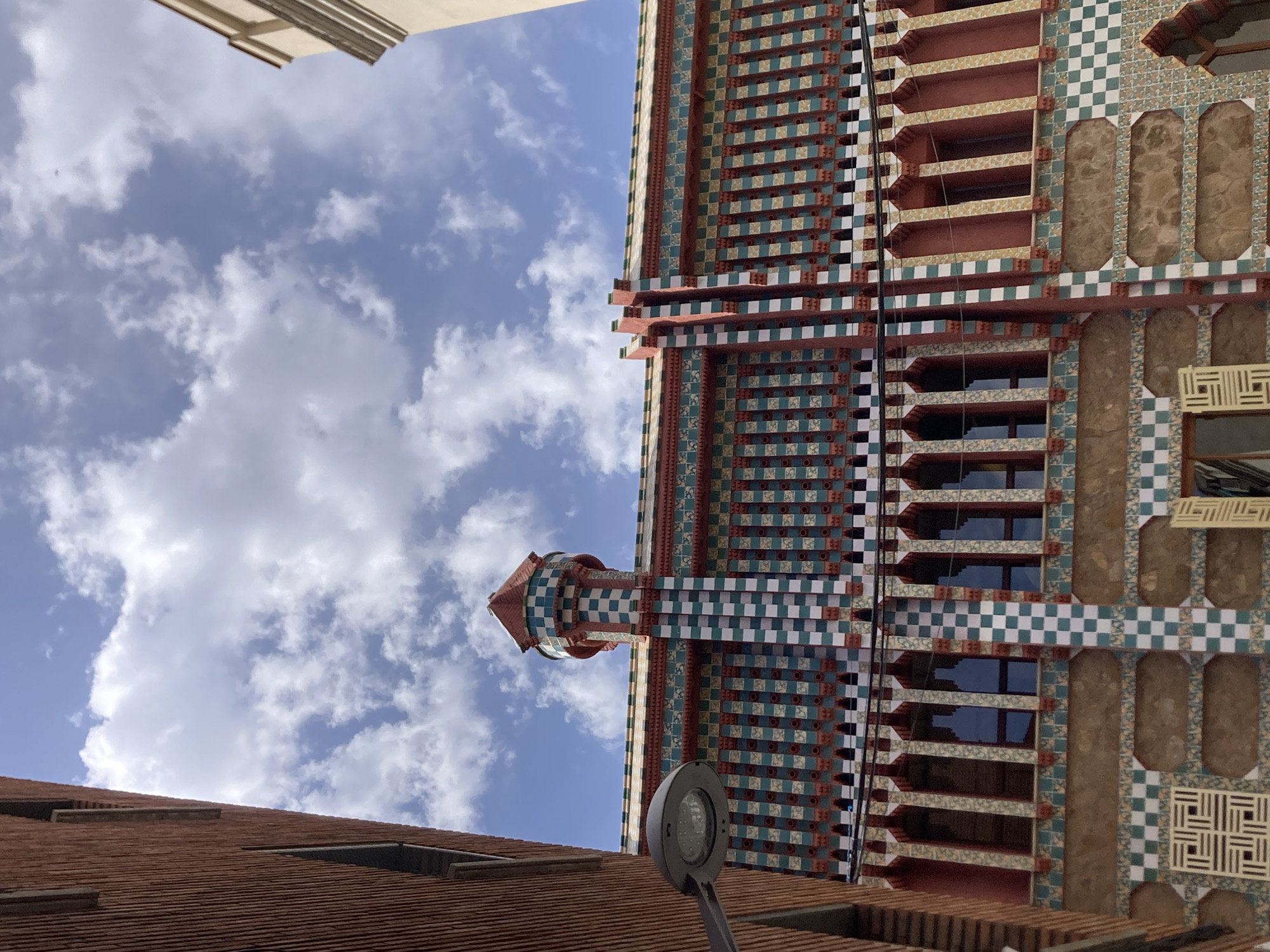 The image shows the facade of Casa Vicens, a building located in Barcelona, Spain. The building is adorned with colorful ceramic tiles and decorative ironwork. The facade features a checkerboard pattern of green and white tiles, interspersed with red and brown accents. Several arched windows are visible, with intricately patterned grilles. A rounded tower with a conical roof, also decorated with tiles, stands prominently on the left side of the building. A modern street lamp is positioned in the foreground on the left. The sky above is a vibrant blue with scattered white clouds. The architectural style is characteristic of Antoni Gaudí's early work, blending Mudejar, Gothic, and Art Nouveau influences.