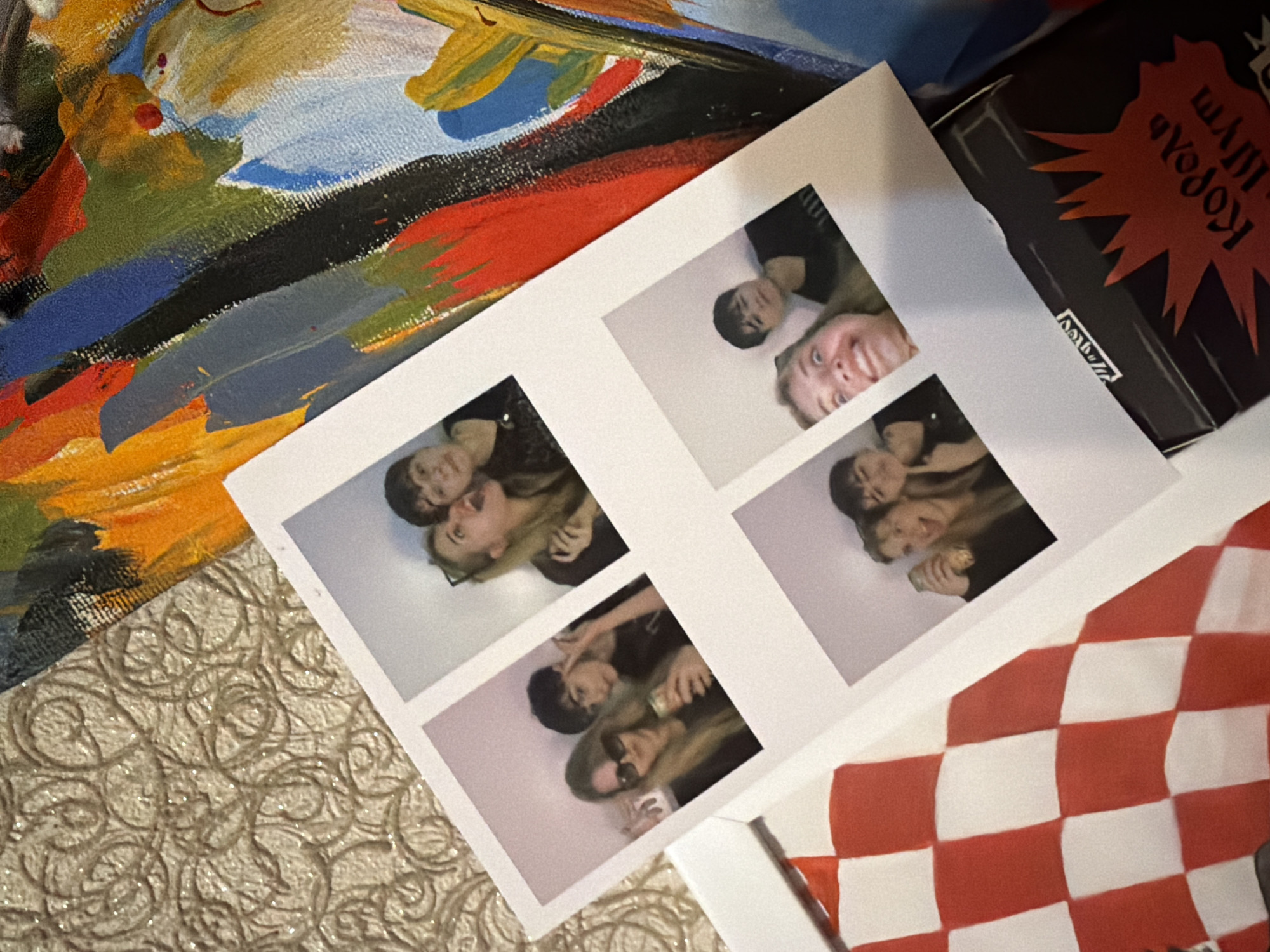 The image shows a collection of items: a canvas with colorful abstract painting in the top-left corner, a black box with Russian text in the top-right corner, a print of four photobooth-style pictures of two people, a red and white checkered object, and a textured surface. The photobooth images appear to show a blonde woman and a dark-haired man in varying poses, potentially together. The surroundings lack context to determine a specific location in Naberezhnye Chelny, Russia.