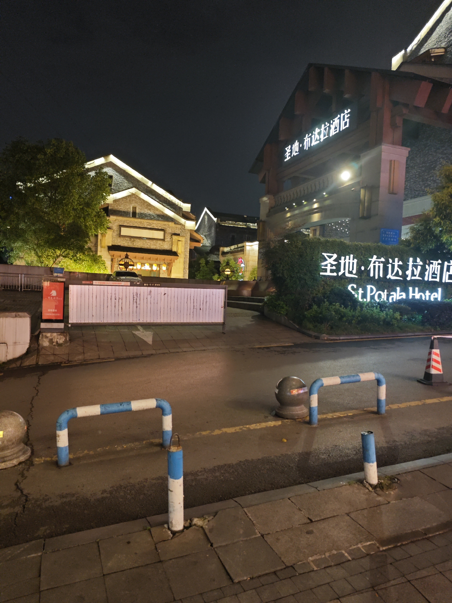 The image captures the St. Potola Hotel in Liangjiang, China, at night. The hotel entrance is visible with signage in both Chinese and English. A gate is positioned across the entrance road, barring access. Roadside bollards painted with blue and white stripes line the foreground of the road. Street lighting illuminates the scene, highlighting the architectural details of the hotel and surrounding buildings. There are no people visible in the image.