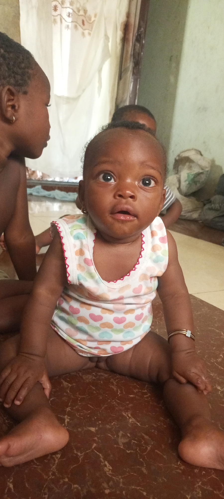 A medium shot depicts a baby seated on a dark brown tile floor, likely indoors. The baby is dark-skinned, wearing a white sleeveless onesie with colorful heart patterns and red trim around the neck and arm openings. The baby has a small gold hoop earring on each ear and a gold and silver bracelet on the right wrist. The baby is looking slightly up and off-center. To the left, partially cropped, is the side of another dark-skinned child with an earring, also seated. Behind the baby is a partial view of another child, and in the background are white curtains. The specific location within Adankolo, Nigeria is not determinable from the image. The floor appears to be brown tile, and behind the baby, there's what may be a painted green wall. In the background is an object that appears to be a pillow on the floor behind the other children.