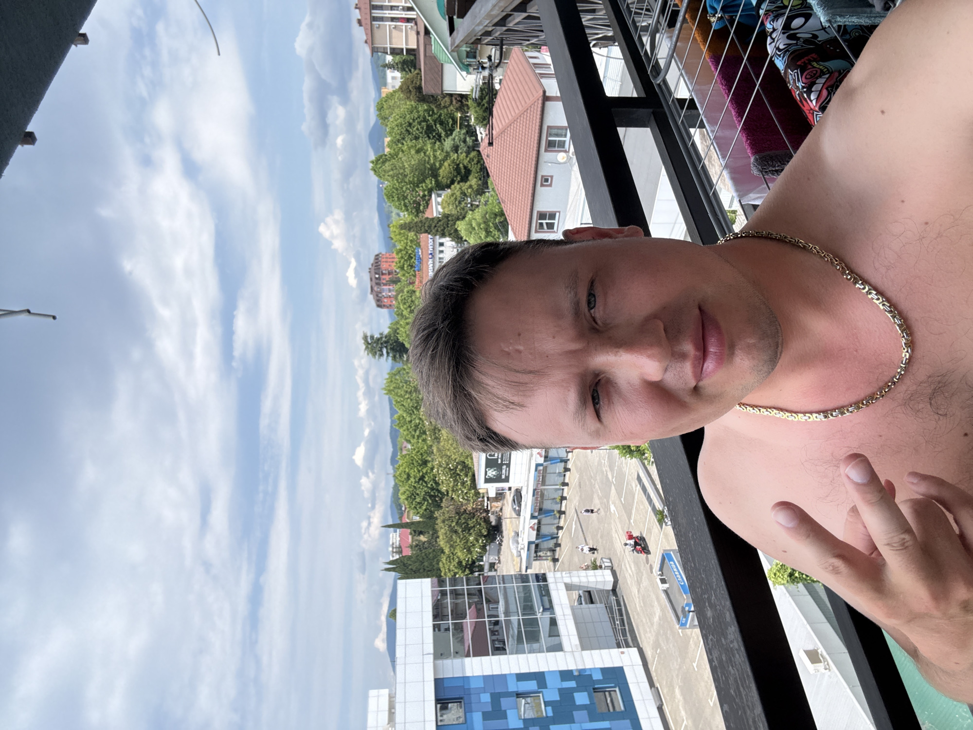 A man is taking a selfie on a balcony in Sochi, Russia. He is wearing a gold chain and making a peace sign with his hand. In the background, there are buildings, trees, and mountains. The sky is cloudy. There is also a clothesline with laundry on it.
