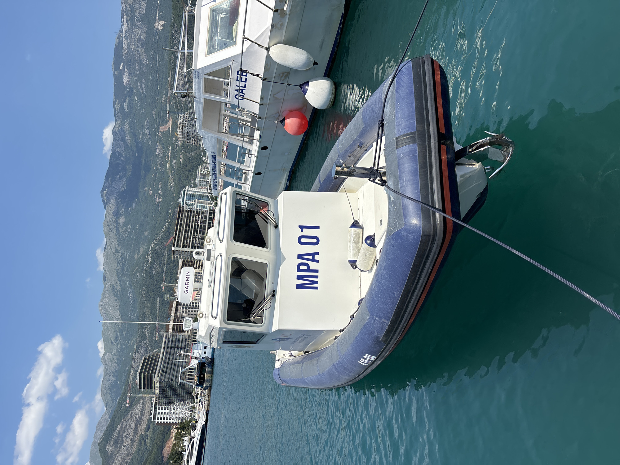 This image depicts a vibrant scene in the marina of Budva, Montenegro, under a clear blue sky with scattered clouds. The time of day appears to be late morning or early afternoon, as indicated by the bright sunlight casting reflections on the water and illuminating the mountainous backdrop.
The primary subjects are two boats docked in the turquoise water. In the foreground, a rigid inflatable boat (RIB) with blue and white pontoons is prominent. The text "MPA 01" is clearly visible in large blue letters on its white cabin. There are also some smaller, less legible markings on the side of the RIB.
Behind the RIB, a larger white boat is partially visible, with the name "GALEB" inscribed on its side. This boat has a cabin with windows and several fenders, including two white oval fenders and a round red one, are attached to its hull, suggesting it is moored.
The background features a picturesque landscape of Budva. Steep, verdant mountains rise behind the marina, and the coastline is dotted with buildings, some of which appear to be under construction, with cranes visible. The water is calm and clear, reflecting the bright sunlight. The overall mood is serene and idyllic, capturing a typical day in a coastal resort town. No people are clearly visible in the image, focusing the attention on the boats and the scenic surroundings. A "GARMIN" logo is visible on a white device on the larger boat.