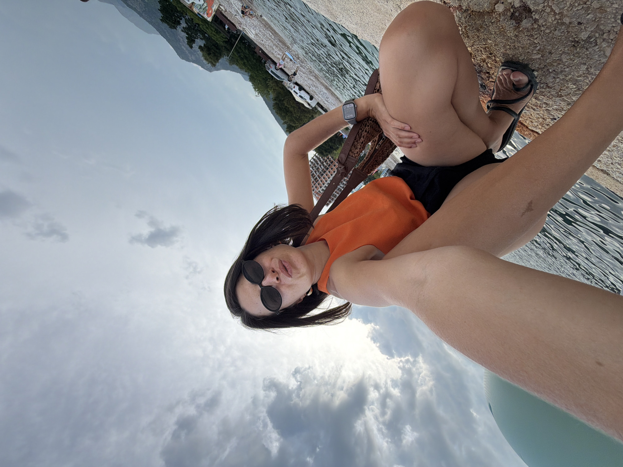 In Budva, Montenegro, a woman is taking a selfie while squatting on a stone pier. She is wearing black sunglasses, an orange sleeveless top, black shorts, and black flip-flops. A brown woven bag is slung over her shoulder. The water of the Adriatic Sea is visible to her right, and a rocky beach with trees and buildings in the background stretches beyond it. The sky above is cloudy.