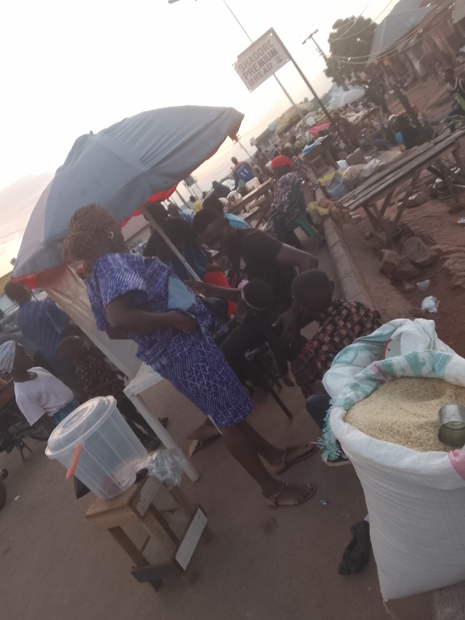 The image depicts a bustling market scene in Gwong, Nigeria. A woman in a patterned purple dress stands near a large sack of grain, likely rice, which is open and filled to the brim. A metal scoop rests on the surface of the grain. Nearby, a man in a black shirt interacts with another person seated in a chair. A child in a checkered shirt stands close to the sack of rice. An umbrella provides shade for the vendor and customers. In the background, there are numerous other people and stalls, suggesting a busy marketplace. A sign for "SHADDORC PREMIUM BREAD" is visible in the distance. The lighting indicates it's either early morning or late afternoon. The environment appears to be a roadside market with wooden tables and other makeshift structures.