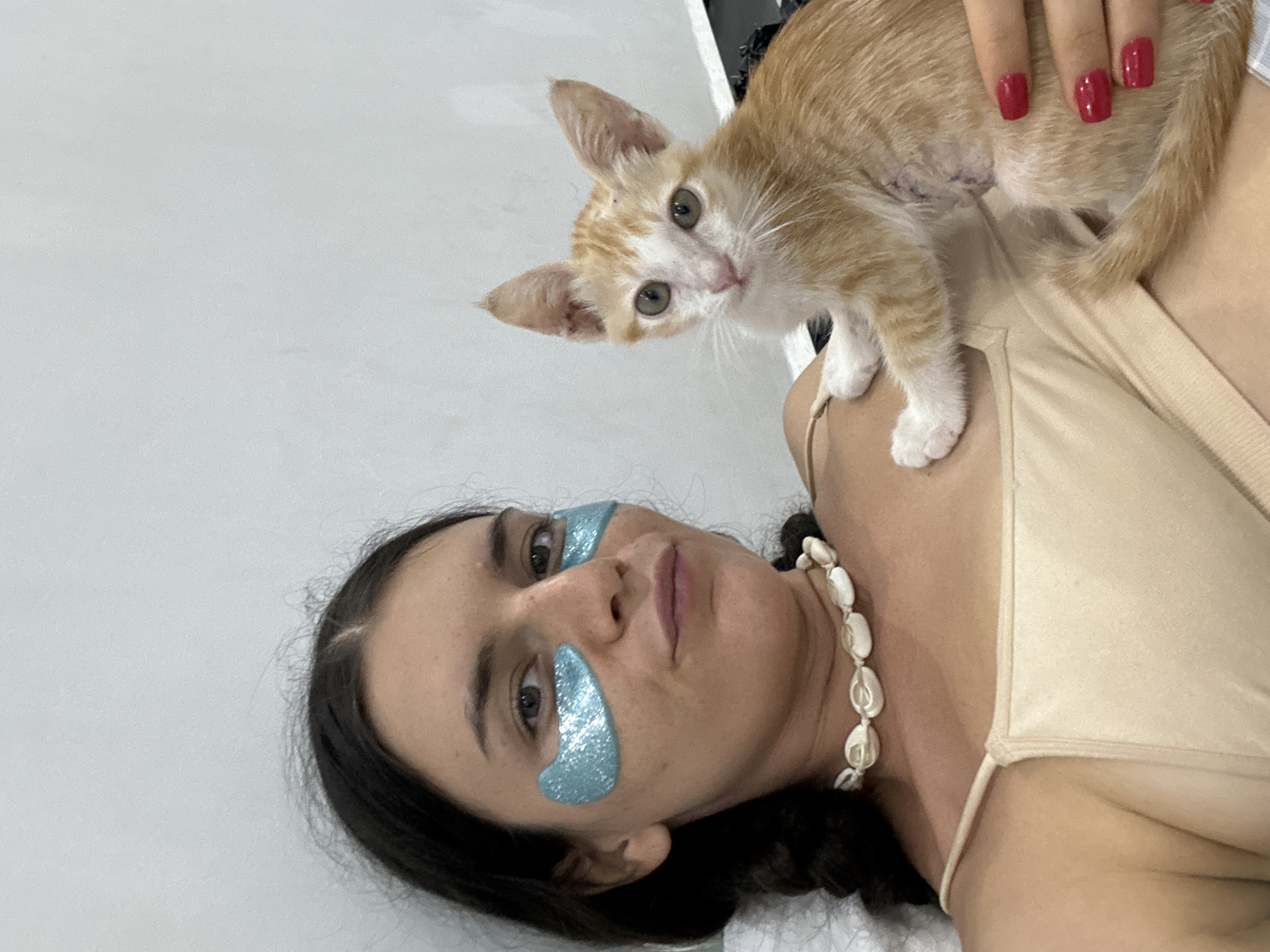 In a close-up, high-angle selfie, a young woman with dark hair styled in braids, wearing a light beige spaghetti strap top and a seashell necklace, poses with a ginger and white kitten. She has glittery blue eye patches on and her fingernails are painted red. The kitten is held gently in her arms, and its fur appears to have some surgical scarring. The background is a plain, light-colored wall. This scene takes place indoors, likely a bedroom or living space, and does not contain any visual elements that definitively place it in Mũi Né, Vietnam.