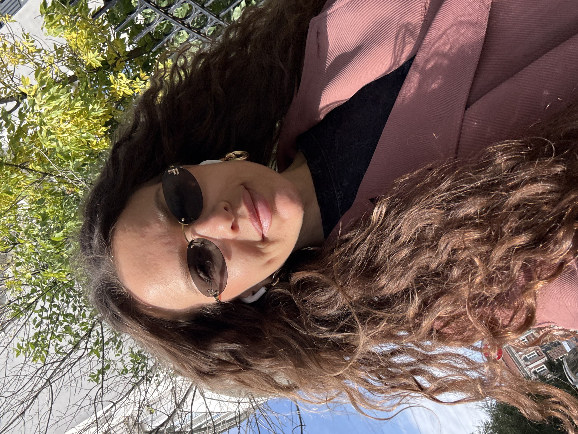 A medium shot, outdoor, eye-level selfie shows a young woman with long, wavy brown hair, wearing sunglasses, a pink blazer over a black t-shirt, and gold hoop earrings. She is also wearing white wireless earbuds. The background consists of trees with green and yellow leaves, a fence, and a clear blue sky with some clouds. The woman appears to be smiling slightly. There is a building with windows visible in the lower left corner, suggesting an urban environment. The image appears to have been taken on a sunny day.