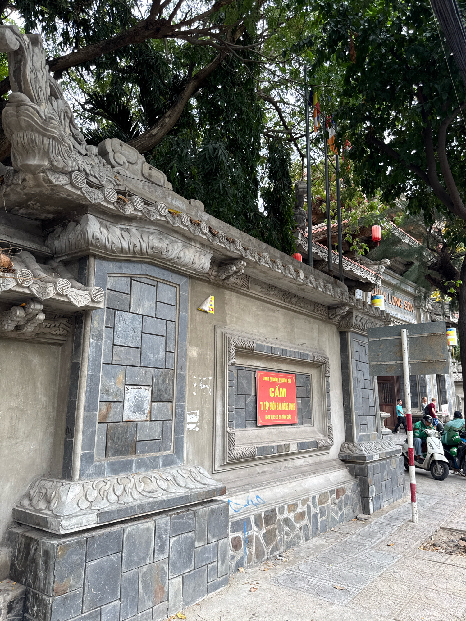 The image captures the entrance to Long Son Pagoda in Nha Trang, Vietnam. A tall, ornate concrete wall, adorned with decorative carvings and grey stone tiles, forms the foreground. On the wall, a red sign with white Vietnamese text reads "CẤM TỰ TẬP BUÔN BÁN HÀNG RONG KHU VỰC CÓ TỔ DÂN PHỐ," which translates to "Prohibited to gather for street vending in the area with the neighborhood group." The wall is topped with intricate, dragon-like sculptures and decorative roof tiles. Behind the wall, the tiled roof of the pagoda is visible, along with several colorful Buddhist flags fluttering in the breeze. Trees with lush green foliage overhang the scene. To the right of the wall, a sidewalk is visible, with a few individuals and a motorcycle parked nearby, suggesting ongoing activity. The overall impression is one of a culturally significant religious site with a bustling urban environment adjacent to it.