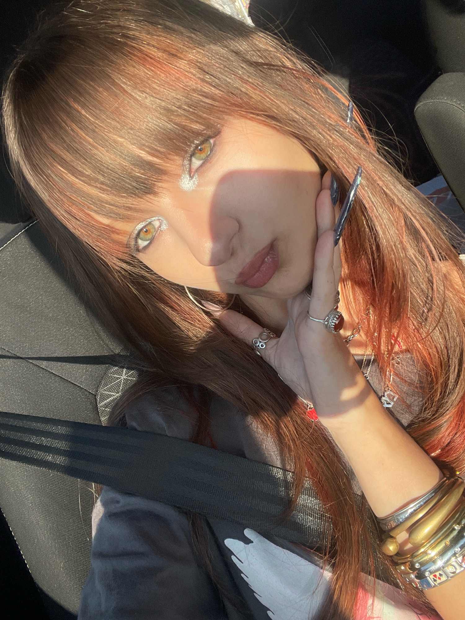 A young woman with long, auburn hair and bangs, wearing a grey sweatshirt, is seen from a close-up, high-angle perspective. She has green eyes with heavy, silver glitter eyeshadow and is wearing multiple rings on her fingers, including one with a large red stone. Her fingernails are long and dark. She is wearing several bracelets on her wrist, including gold and silver bangles. The woman is inside a car, as indicated by the seatbelt and car seat visible in the frame. The sun is shining brightly, casting a shadow across her face and hair. The image does not contain enough visual information to determine a specific location in the city of Raanana, Israel.