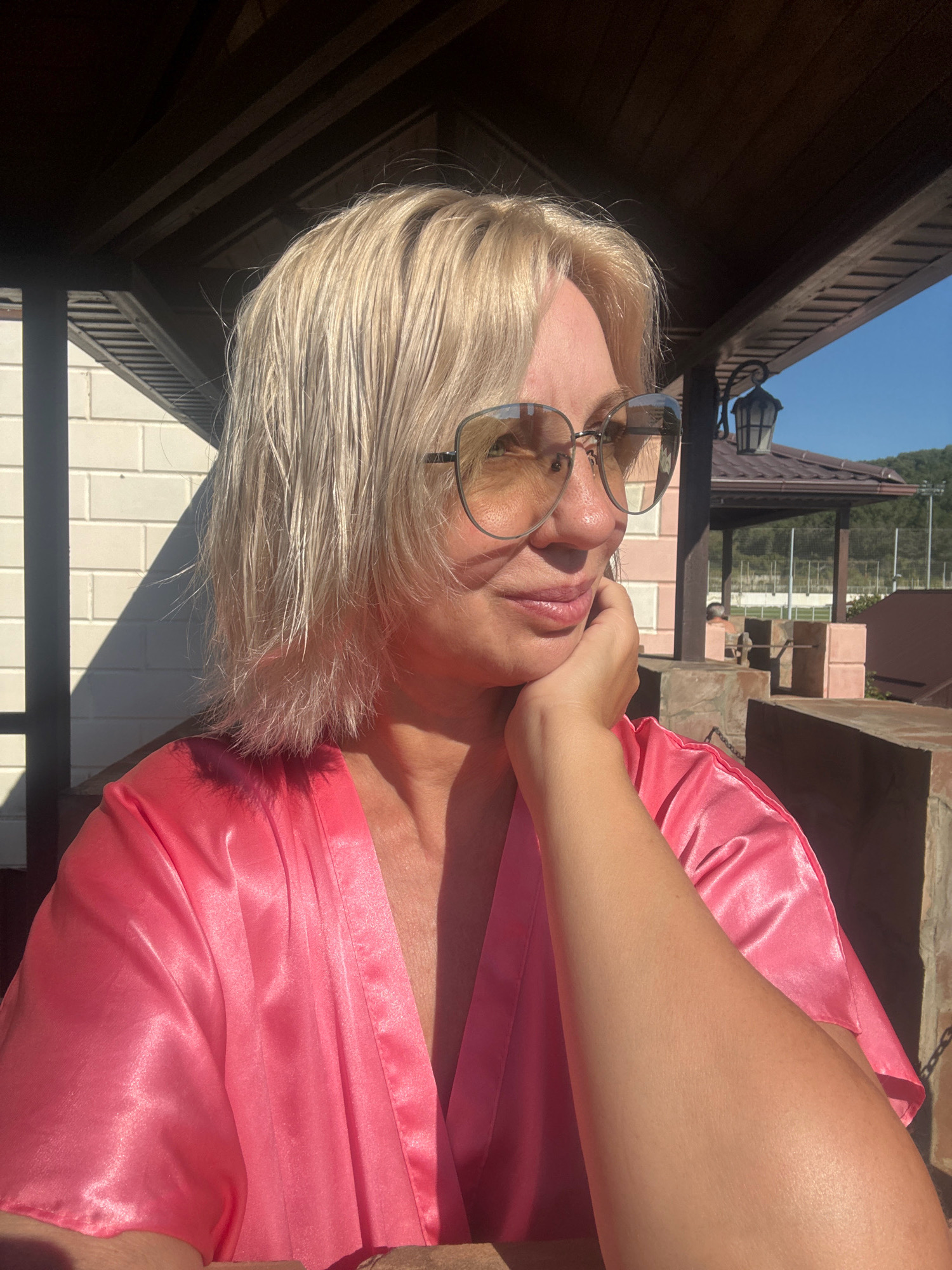 A woman with blonde hair and sunglasses is sitting outside on a sunny day. She is wearing a pink satin robe. The background suggests an outdoor setting, possibly a resort or leisure area, with a tiled roof structure and some greenery visible. There's a shadow cast on a white brick wall behind her. The overall impression is one of relaxation and leisure. The image does not provide specific clues to pinpoint the location within Abrau-Dyurso, Russia, beyond it being an outdoor, likely recreational, setting.