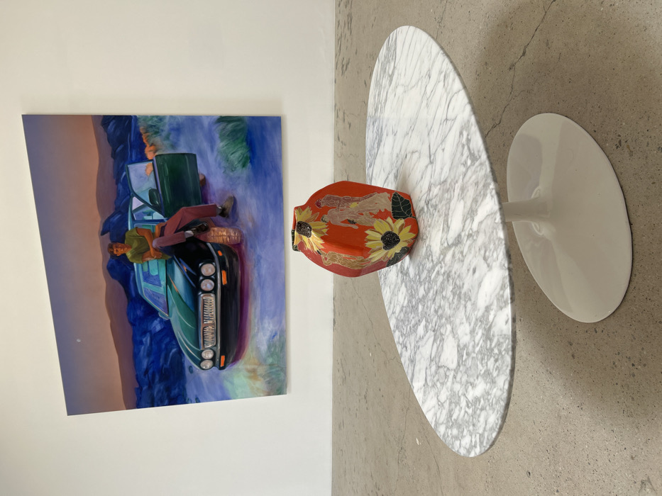 A painting depicts a man leaning on a car on a roadside, with blue and purple hues surrounding them. Next to the painting, on a marble-patterned round table with a white base, sits an orange vessel adorned with sunflowers. The table and the vessel are placed on a light gray floor.