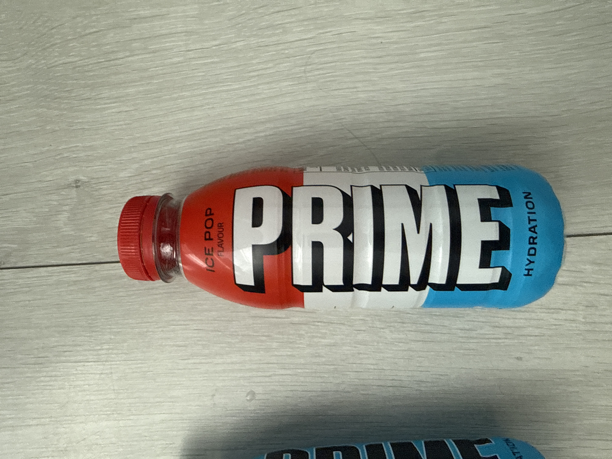A single bottle of Prime Hydration, Ice Pop flavor, is pictured standing upright on a light-colored wooden surface. The bottle has a red cap and a label that is predominantly red, white, and blue. The word "PRIME" is written vertically in large white letters with black outlines. Above "PRIME," "ICE POP FLAVOUR" is printed in smaller black text on a red background. Below "PRIME," the word "HYDRATION" is printed in black text on a blue background. The bottle is centered in the frame. The background is a light-colored wooden surface with visible grain lines, likely a floor or table. The lighting is even and does not create harsh shadows. No people, animals, or other objects are visible in the image. The location is not specified in the image, so a precise location in Burgas, Bulgaria cannot be determined.