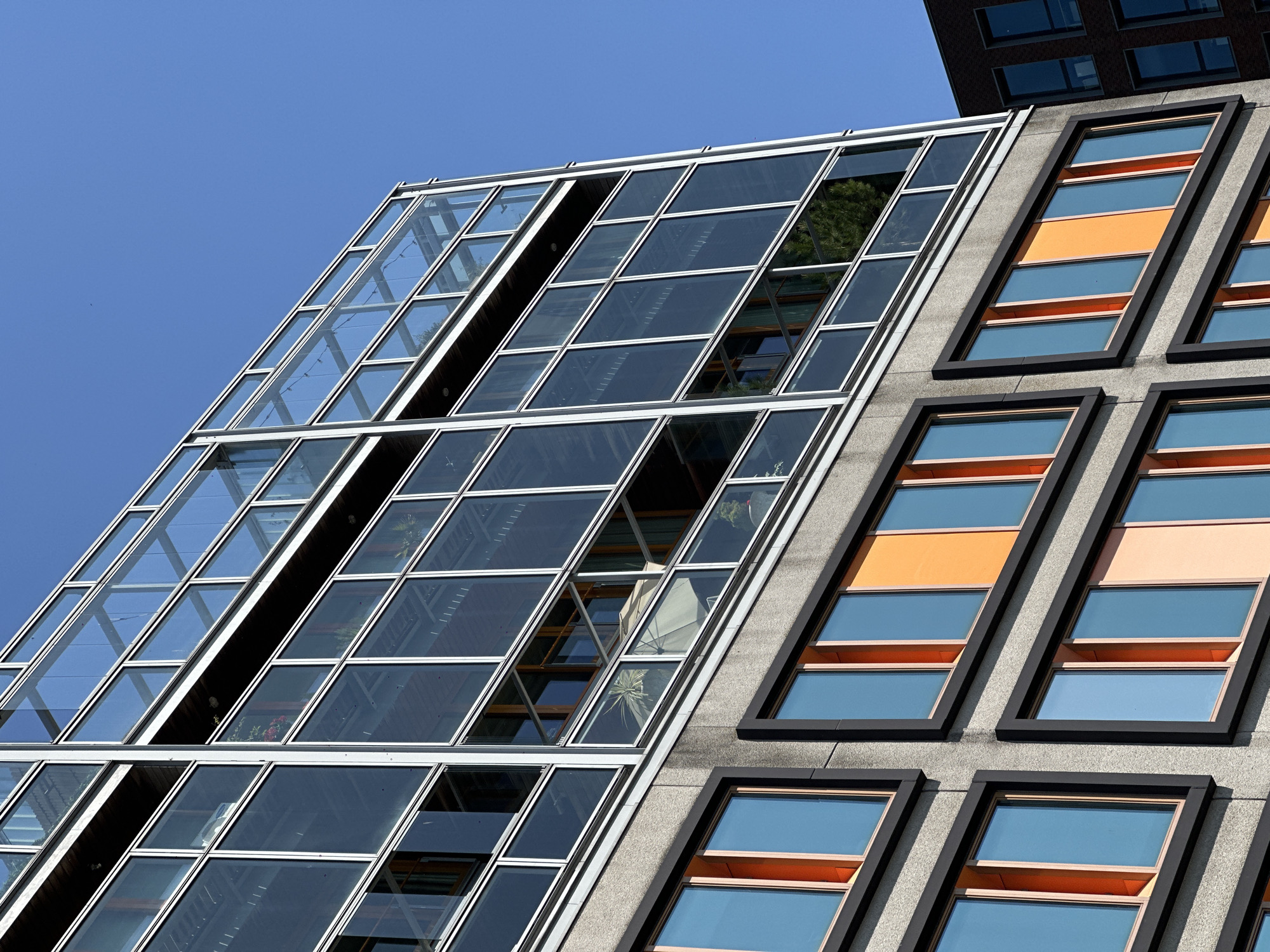 The image depicts a section of modern architecture, showing two distinct building facades. The left side features a glass-enclosed structure with a grid pattern, with some of the windows slightly ajar. The right side presents a concrete facade with rectangular windows framed in a dark color, some containing orange panels. The windows reflect the blue sky. The overall composition is taken from a low-angle perspective, emphasizing the height of the buildings. The location is likely Amsterdam, Netherlands, due to the characteristic architectural style.