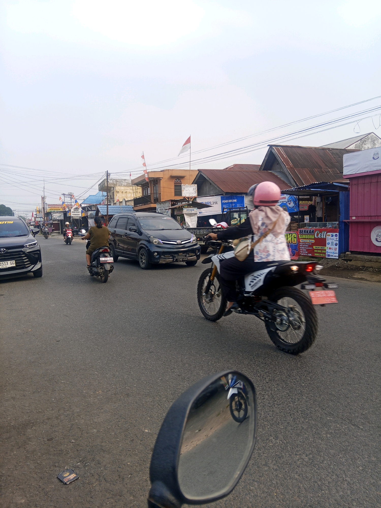 The image captures a street scene in Lubuklinggau, Indonesia, featuring various vehicles and buildings. In the foreground, a motorcycle is being ridden, with the rider wearing a pink helmet and a patterned shirt, and a passenger behind them, also wearing a helmet. To the left, a black SUV is driving away, and another motorcycle is visible in the distance. Further down the street, other vehicles and buildings line the road, including shops with various signs and awnings. Indonesian flags are displayed on some of the buildings. The sky is overcast.