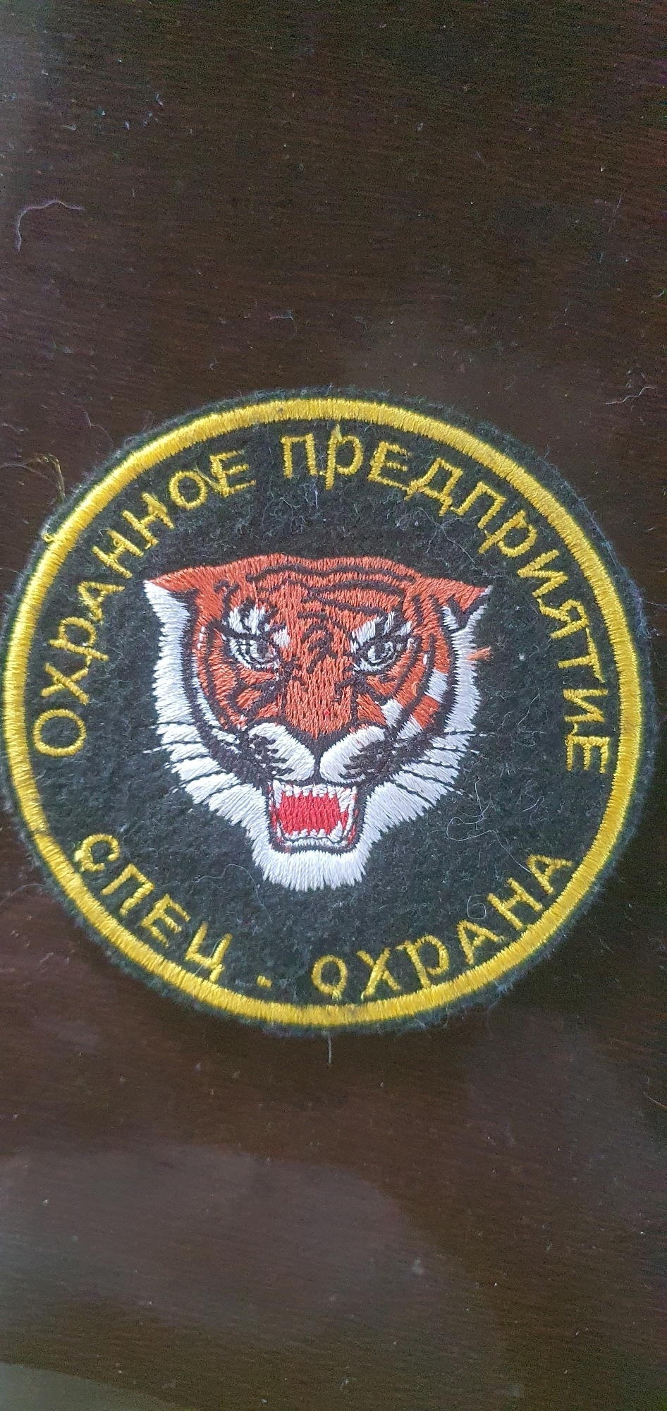 A round, embroidered patch is centered on a dark, textured surface, likely wood. The patch features a yellow border with text in yellow Cyrillic letters. The upper arc of the text reads "ОХРАННОЕ ПРЕДПРИЯТИЕ" and the lower arc reads "СПЕЦ - ОХРАНА". Within the border, the head of a tiger is depicted. The tiger has orange fur with black stripes, white markings around its eyes and snout, and a fierce expression with bared teeth. The background of the patch is black. The surface beneath the patch is dark brown with a visible wood grain and some faint lighter specks and smudges. The lighting appears to be from above, casting a subtle shadow to the left of the patch. There is no indication of the city of Chastoe, Russia, within the image itself.