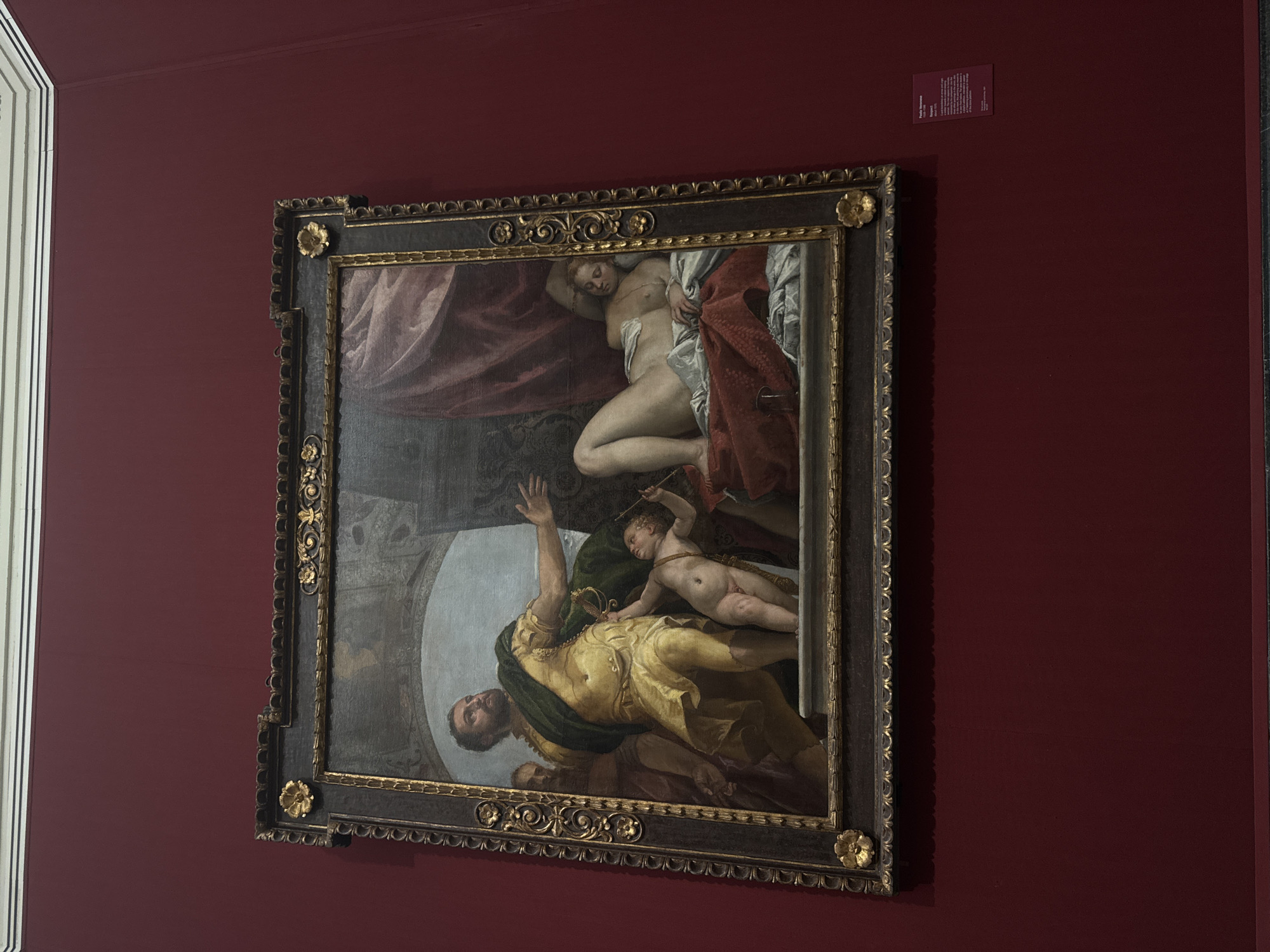 The image shows a painting in a museum. The painting is framed with an ornate, dark brown and gold frame, and it is displayed on a deep red wall. The painting depicts a scene with several figures. On the left, a man in a yellow and green tunic stands and gestures with his right hand. Next to him, a cherubic figure, possibly a cupid, reaches up towards the man's hand. To the right of the man, a woman reclines on a red and blue draped surface, her body partially visible. Behind her, heavy red drapery fills the upper right portion of the painting. The background of the painting shows architectural elements, including an archway. A small, rectangular information plaque is visible on the wall to the right of the painting. The painting and its display suggest it is an artwork of historical or artistic significance, likely in a public gallery. The environment is a museum gallery, identified by the wall color, lighting, and display of artwork. The overall scene is a static depiction of a posed tableau within a painting, presented in a museum setting. Based on the provided crops which reveal text on a label, the painting is by Paolo Veronese, titled "Respiro," and dates from 1575-1578. This suggests the artwork is likely housed in a major art institution in London, United Kingdom, such as The National Gallery.