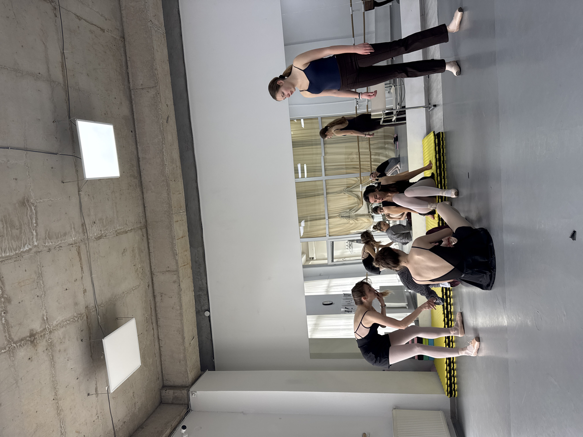 The image captures an indoor ballet studio, likely located in Tbilisi, Georgia. Several young women, presumably dancers, are visible, engaged in a practice or training session. In the foreground, one woman stands, wearing a navy top and dark pants, with her hair pulled back. Further into the studio, a group of dancers is seated or stretching on the floor. Most are dressed in black leotards and light-colored tights, with some wearing pointe shoes. Their figures are reflected in a large mirror that spans a significant portion of one wall, also showing reflections of ballet barres and windows.
The studio's architecture features a raw, industrial aesthetic with an exposed concrete ceiling and pillars, contrasting with the smooth, light gray floor and clean white walls. Two rectangular fluorescent light panels are suspended from the concrete ceiling, illuminating the space. Yellow and black interlocking foam mats are laid out on the floor, suggesting an area for floor exercises or stretching. Small white security cameras are mounted on the walls. The overall scene suggests a dedicated space for dance training. No visible text is clearly discernible.