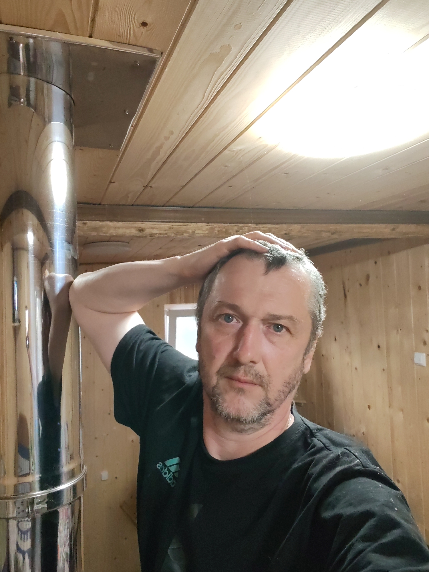 The image shows a man with short grey hair and a beard, wearing a black t-shirt with the Adidas logo. He has his left arm raised, with his hand resting on his head, and he is looking towards the camera. The man appears to be indoors, in a room with wooden paneling on the walls and ceiling. A large, shiny metal pipe, likely a chimney flue, is visible on the left side of the frame, extending upwards. The lighting suggests it might be daytime, with a bright overhead light fixture visible on the ceiling. The overall impression is casual and informal, possibly a selfie taken in a home or workshop environment. The scene is likely set in Chaikovsky, Russia, based on the provided location context.