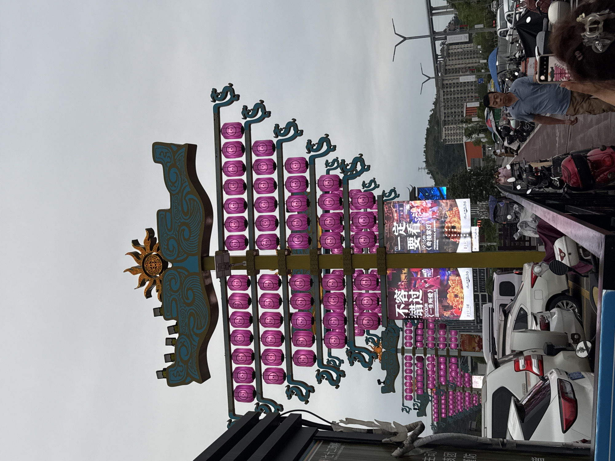 The image depicts a complex street scene in Zhangjiajie, China. A prominent decorative sign, featuring stylized patterns, a sun emblem, and multiple rows of purple lanterns, dominates the left side of the frame. Adjacent to the sign are illuminated advertising displays, partially obscured by parked white vehicles. A man, captured mid-selfie, stands near parked motorcycles on the right side of the image. In the background, buildings, a bridge structure, and wind turbines are visible under an overcast sky. Additional signs and street-level details are present but partially obscured by the overall composition.