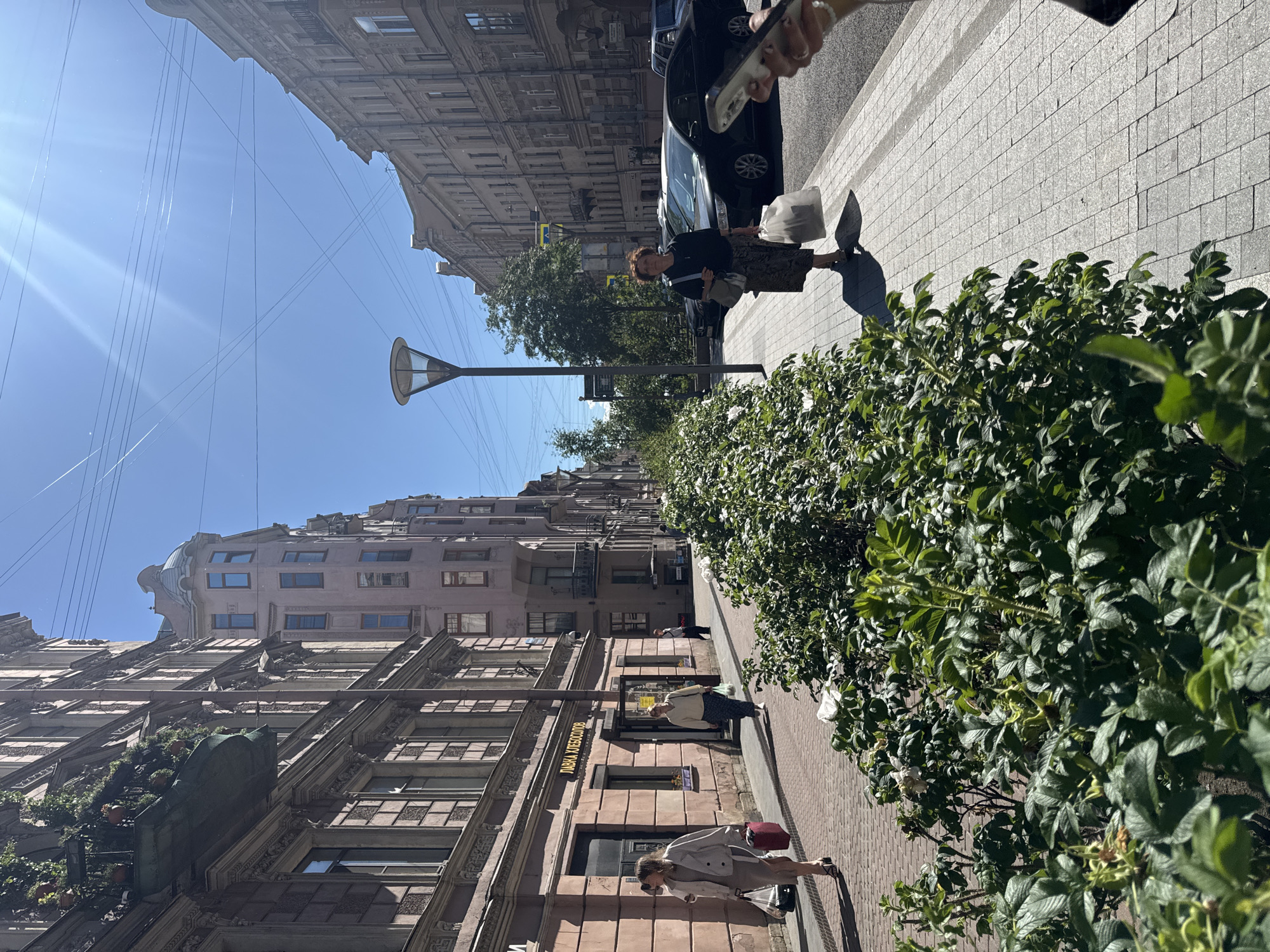 The image depicts a street scene in Saint Petersburg, Russia, on a sunny day, likely in the late morning or early afternoon, given the strong sunlight and sharp shadows. The perspective is angled, looking down a street lined with ornate, multi-story buildings characteristic of the city's architecture. Lush green bushes and trees are visible in the foreground and along the sidewalk, adding a touch of nature to the urban environment.
Several people are present on the sidewalk. In the mid-ground, a woman with short, reddish-brown hair is walking, carrying a white bag and a light-colored purse. Further down the street, another woman is walking with a red handbag, wearing a light jacket and sunglasses. Closer to the foreground, partially obscured by foliage, another woman is seen walking. A black car is parked on the street.
Overhead, a street lamp with a modern design is visible, along with numerous electrical wires crisscrossing the bright blue sky. The buildings themselves exhibit detailed facades with numerous windows, balconies, and decorative architectural elements. One building has a sign with Cyrillic text: "ЛАВКА ХЛЕБОСОЛОВ" which translates to "Lavka Khlebosolov" (a type of shop often selling bread and other provisions). The overall atmosphere appears to be one of everyday life on a pleasant day.
