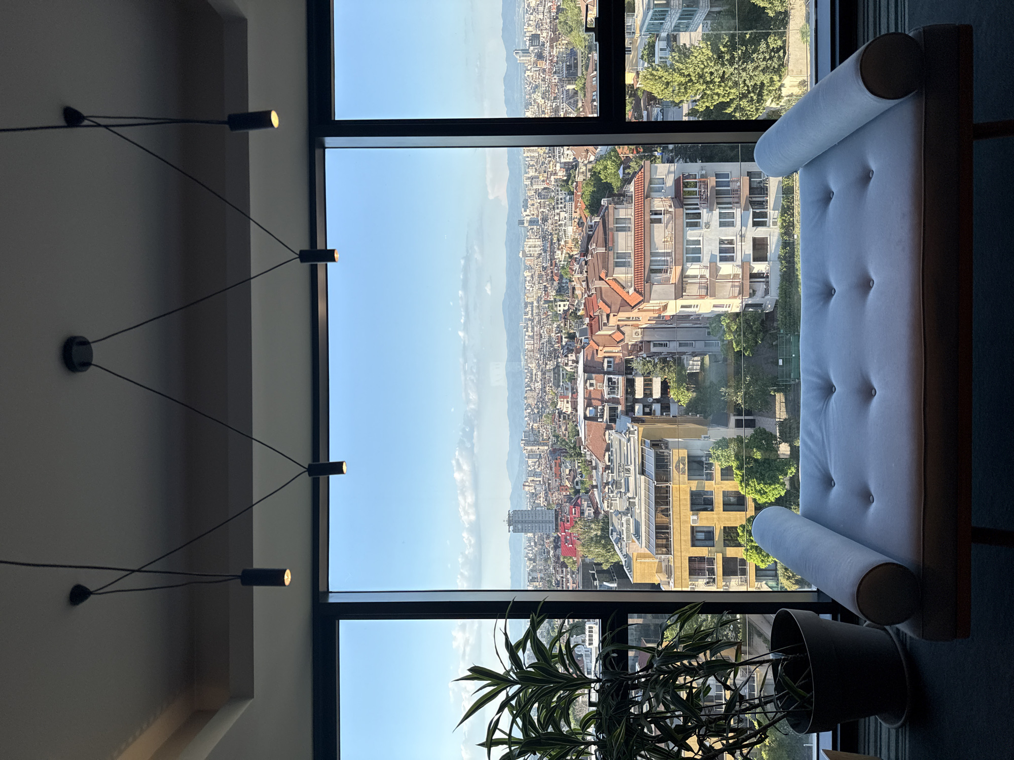 A high-angle, interior view shows a city scene through a multi-pane window in Sofia, Bulgaria. The city consists of densely-packed buildings, mostly of low to mid-rise construction, extending towards distant hills under a clear sky with some clouds.
Inside, on the right side of the frame, is a light-colored chaise lounge with cylindrical headrests and a buttoned surface. At the bottom of the frame and partially obstructing the view is a potted plant with long, green, leafy fronds. On the left side, a modern, multi-armed light fixture is affixed to the wall. The window frames are black.