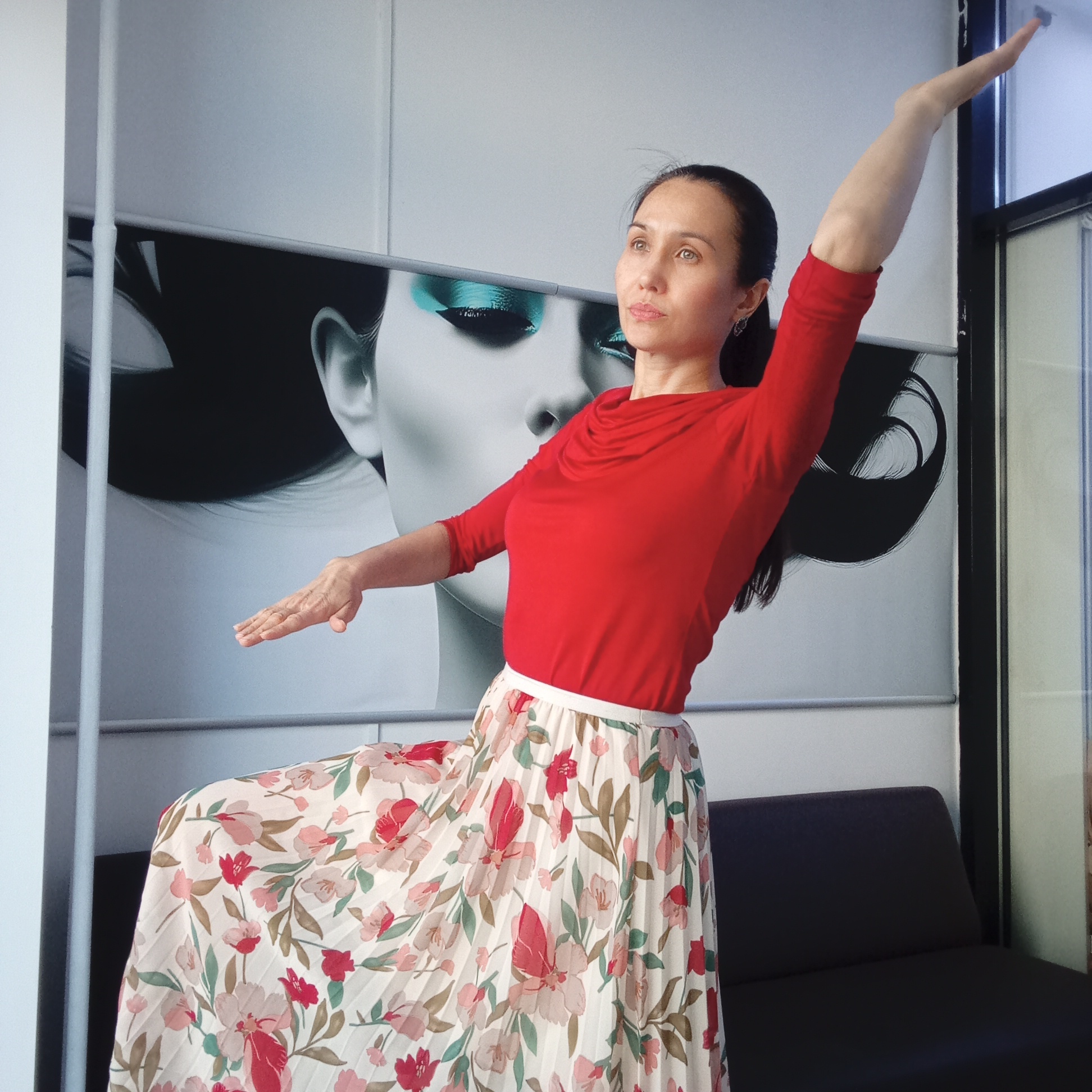A full-length photograph depicts a woman in a red, long-sleeved top and a long, floral skirt striking a dance pose. Her right arm is raised high and extended, while her left arm is extended to the side at shoulder level. The woman has dark hair pulled back and is looking to the right.
Behind her is a wall with a large, black and white advertisement featuring a woman's face with bright blue eye makeup. To the right of the advertisement is a black framed window. In the lower right of the frame, the top of a black couch is visible.
Based on visual evidence and research, the location appears to be a dance studio or similar space located in Izhevsk, Russia. However, the image lacks specific geographical indicators to confirm the exact location.
