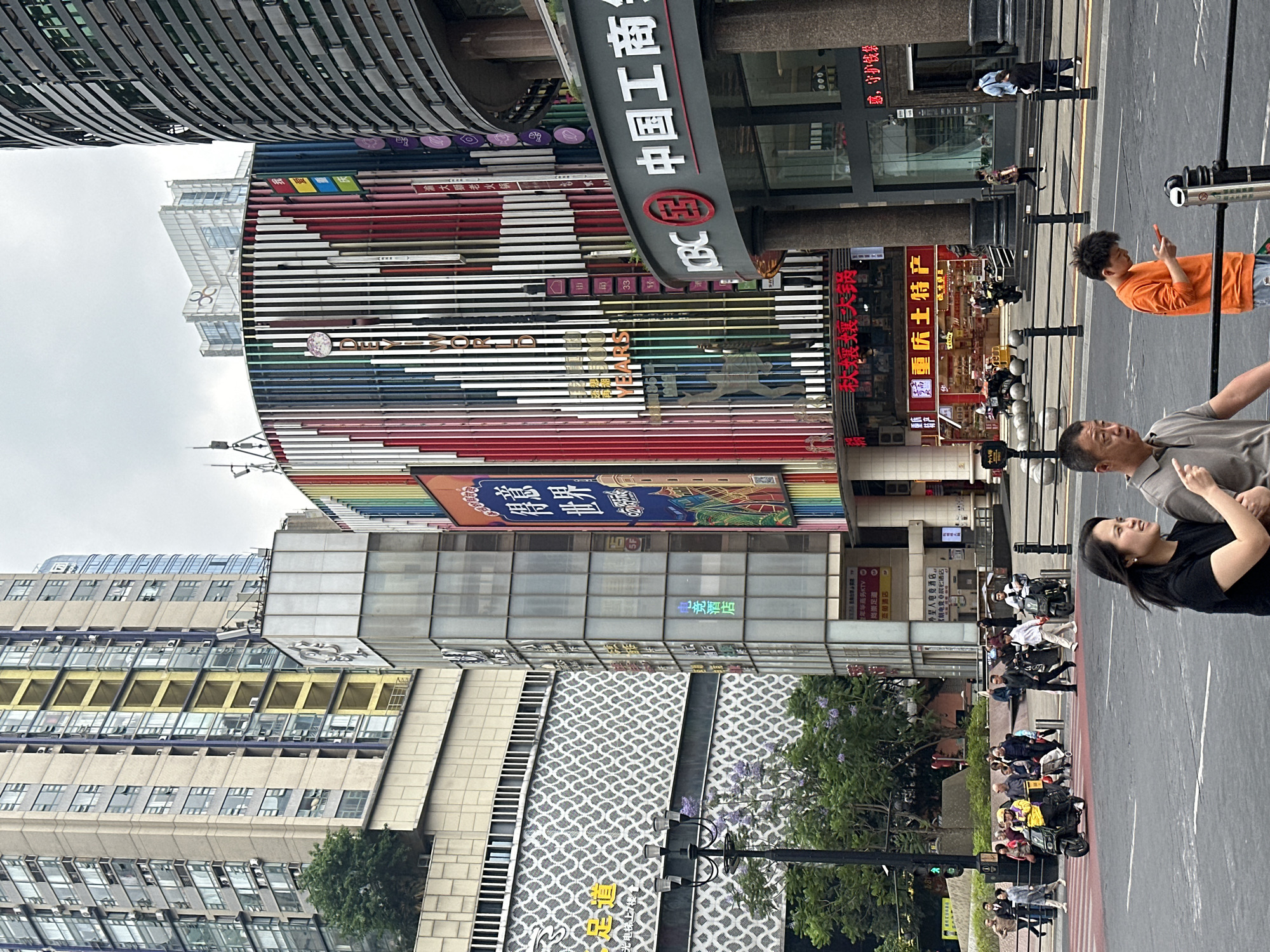 The image shows a bustling urban street scene in what appears to be the Yuzhong District, China, recognizable by the presence of Chinese signage and architecture. In the foreground, several people stand on the sidewalk. A woman in a black shirt is looking upwards, possibly in conversation with a man beside her. A boy in an orange shirt is also visible, with his hand raised in a pointing gesture.
The buildings in the background are tall and densely packed, featuring a mix of architectural styles, including a building with a curved facade adorned with horizontal stripes of various colors. Various advertisements and signage are visible on the buildings, indicating different businesses and establishments. Street signs such as "中国工商" and store signs such as "重庆土特产" are displayed, identifying specific locations.
The street is busy with pedestrian traffic, and there are vehicles visible in the distance. The overall environment suggests a vibrant commercial area in a major city.