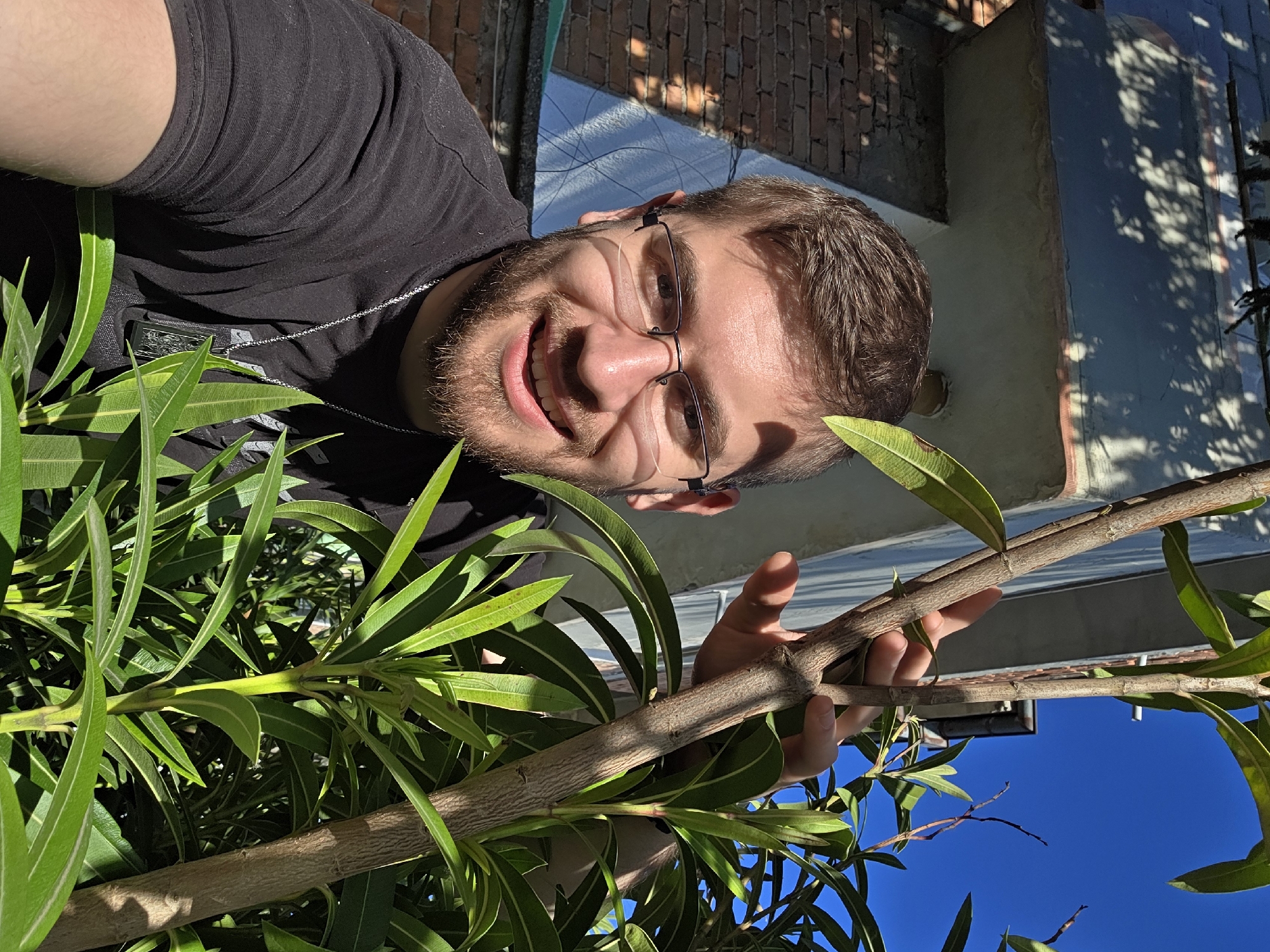 A man with a beard and glasses, wearing a black shirt and a necklace, smiles at the camera. He is positioned low, looking up towards the camera, holding a leafy tree branch. The clear blue sky is visible in the lower right corner of the frame. A brick building is in the background. Given the prompt including Yagodovo, Bulgaria, this photo was most likely taken in or around Yagodovo, Bulgaria.