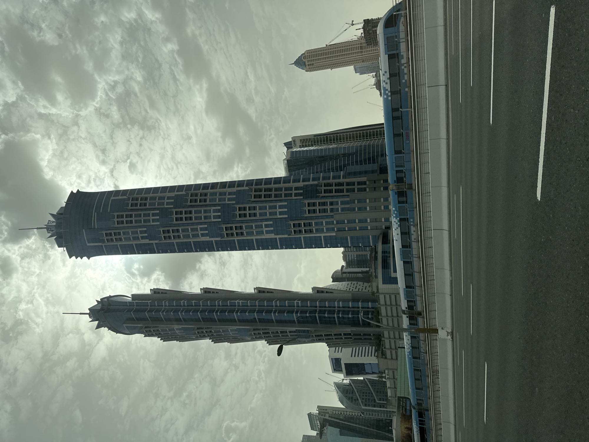The image shows a view of Business Bay in Dubai, United Arab Emirates. The view is from a bridge or elevated road, with the road surface and lane markings visible in the foreground. Buildings are visible to the left. The sky is overcast with heavy clouds.