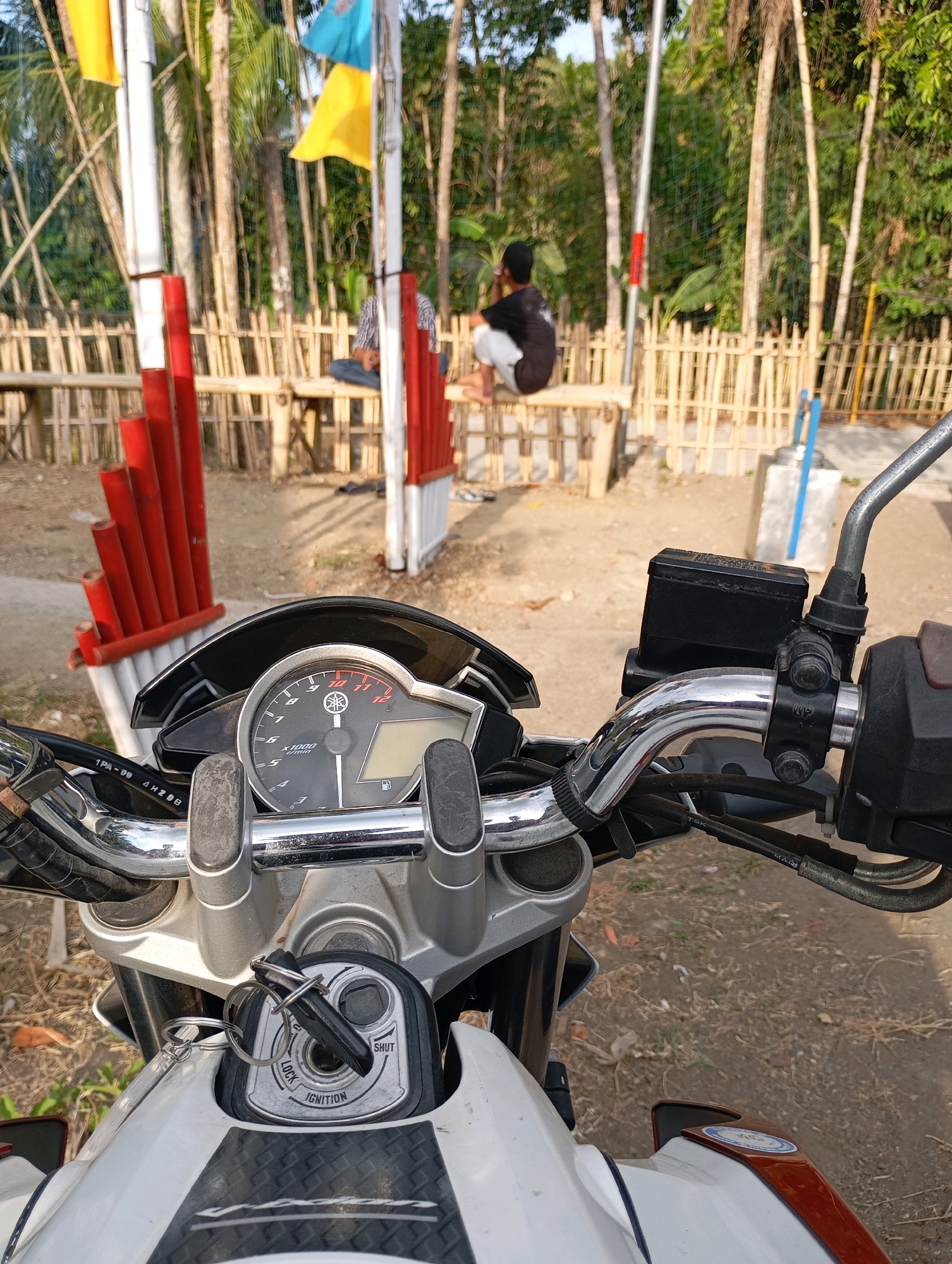 The image shows a close-up view of a motorcycle's handlebars and ignition from the perspective of someone sitting on the motorcycle. In the background, there is a bamboo fence and several people sitting on a bamboo bench. There are also flags on poles in the background, and trees and foliage suggesting an outdoor, possibly rural, environment. The scene appears to be in the city of Cigunung, Indonesia, given the context provided. The motorcycle is white and has a visible speedometer and ignition key. The people in the background are not clearly identifiable. The overall impression is a casual outdoor setting with a focus on the motorcycle.