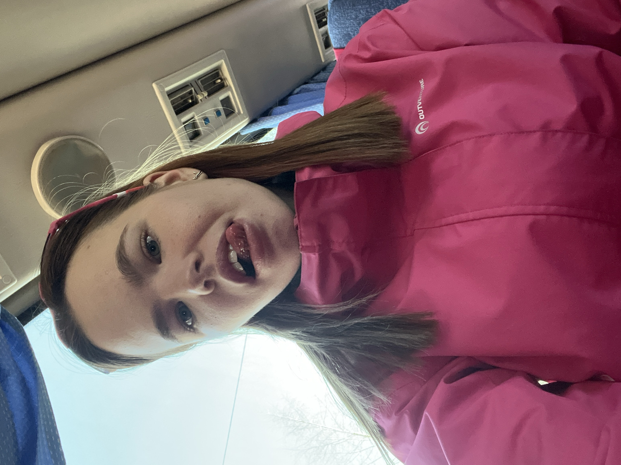 A young, fair-skinned individual with light eyes and light brown hair, wearing a pink winter jacket, is positioned inside a vehicle, possibly a bus or van, as indicated by overhead vents and speaker. The person is sticking out their tongue and looking directly at the camera. The environment outside the vehicle appears overcast, with bare trees visible through the window, suggesting it may be late fall or winter. The location cannot be determined based on the provided visual information. There is no indication it is located in Berezniki, Russia.
