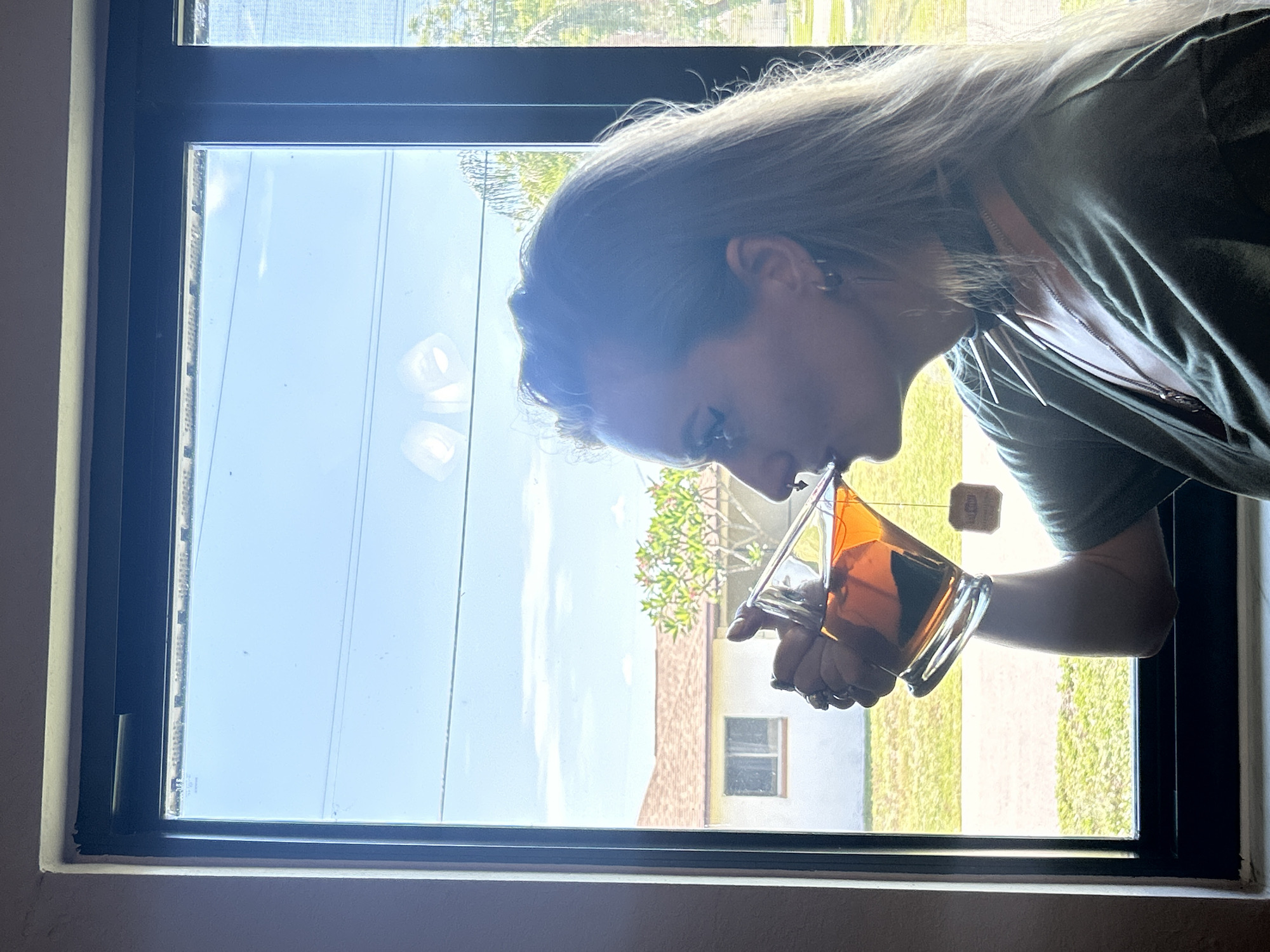 A woman is seen in profile, holding a glass of iced tea and drinking it. She has long, blonde hair and is wearing a green shirt and a choker necklace. The scene takes place indoors, in front of a window that looks out onto a residential area in Cape Coral, Florida. The background shows a house with a tiled roof, a small tree, and power lines against a clear blue sky with some clouds. The lighting suggests it is daytime.