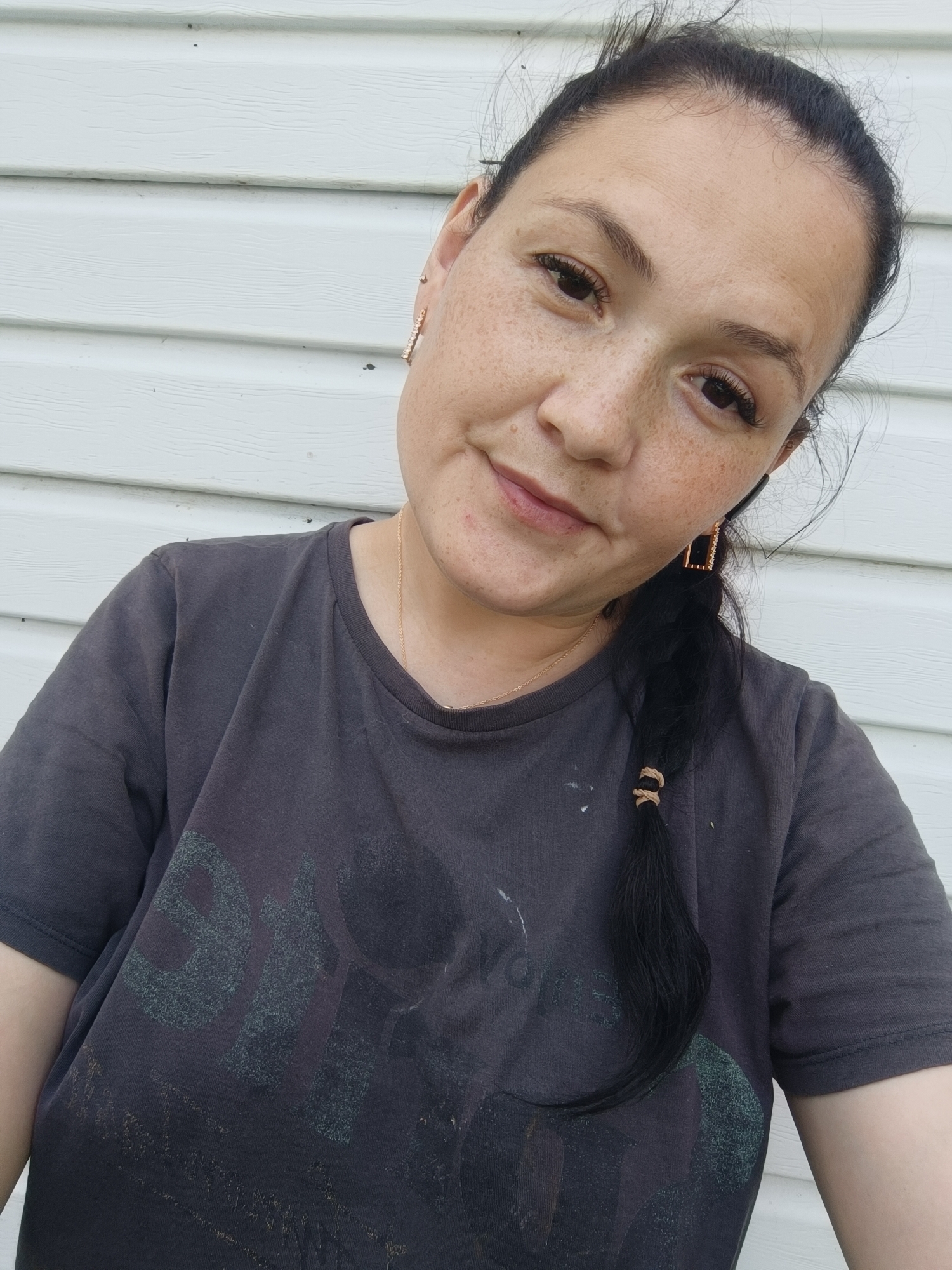 A woman with dark hair pulled into a braid is leaning into the camera, a slight smile on her face. She is wearing a dark grey t-shirt and several pieces of jewelry, including gold earrings and a thin gold necklace. The background is a white, horizontally-paneled wall. The image is a selfie.