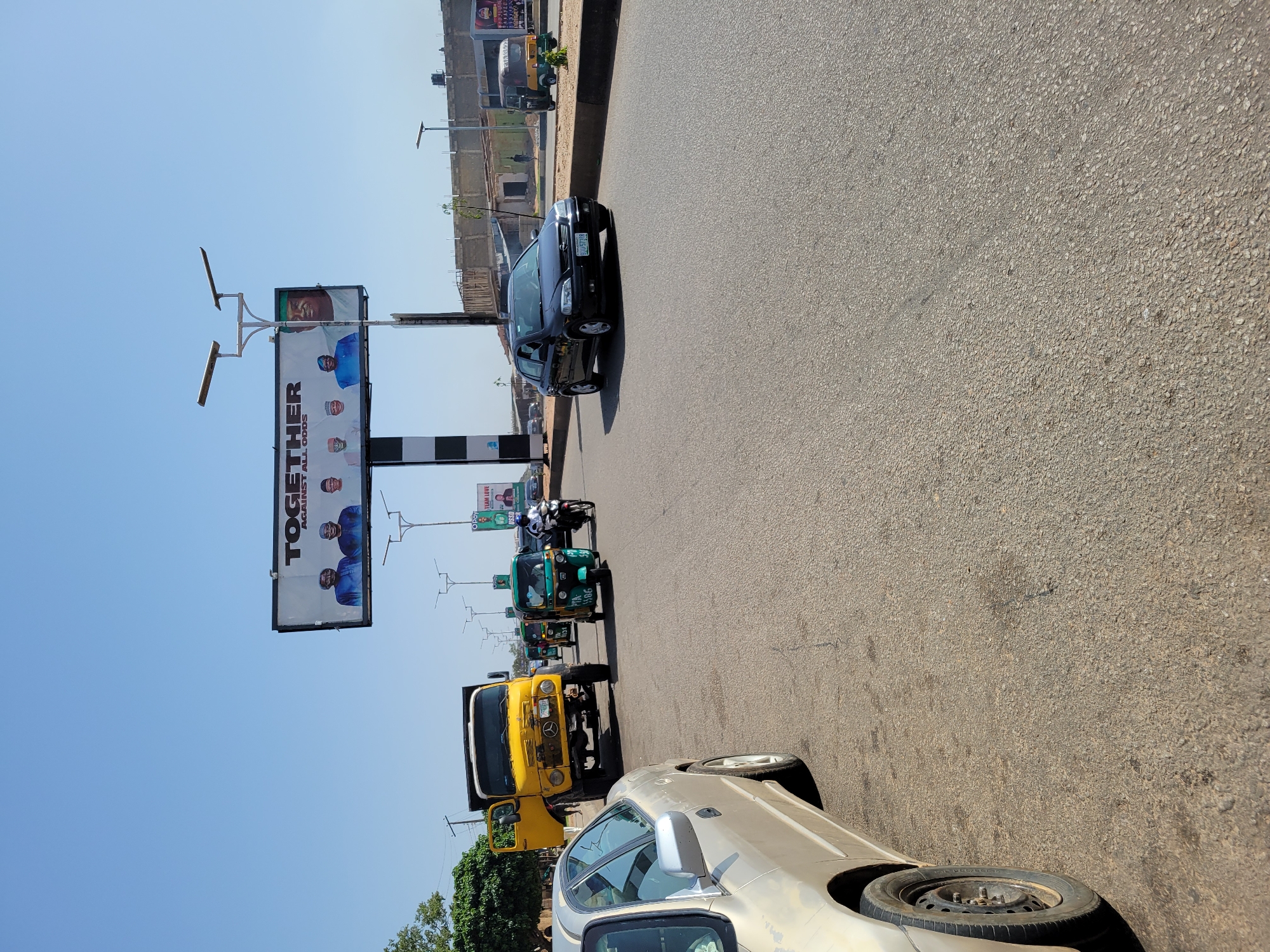 The image shows a street in Unknown, Unknown on a clear day. A large billboard with the word "TOGETHER" is visible. A mix of vehicles, including cars, a yellow truck, and green auto-rickshaws are on the road. The road surface is asphalt.