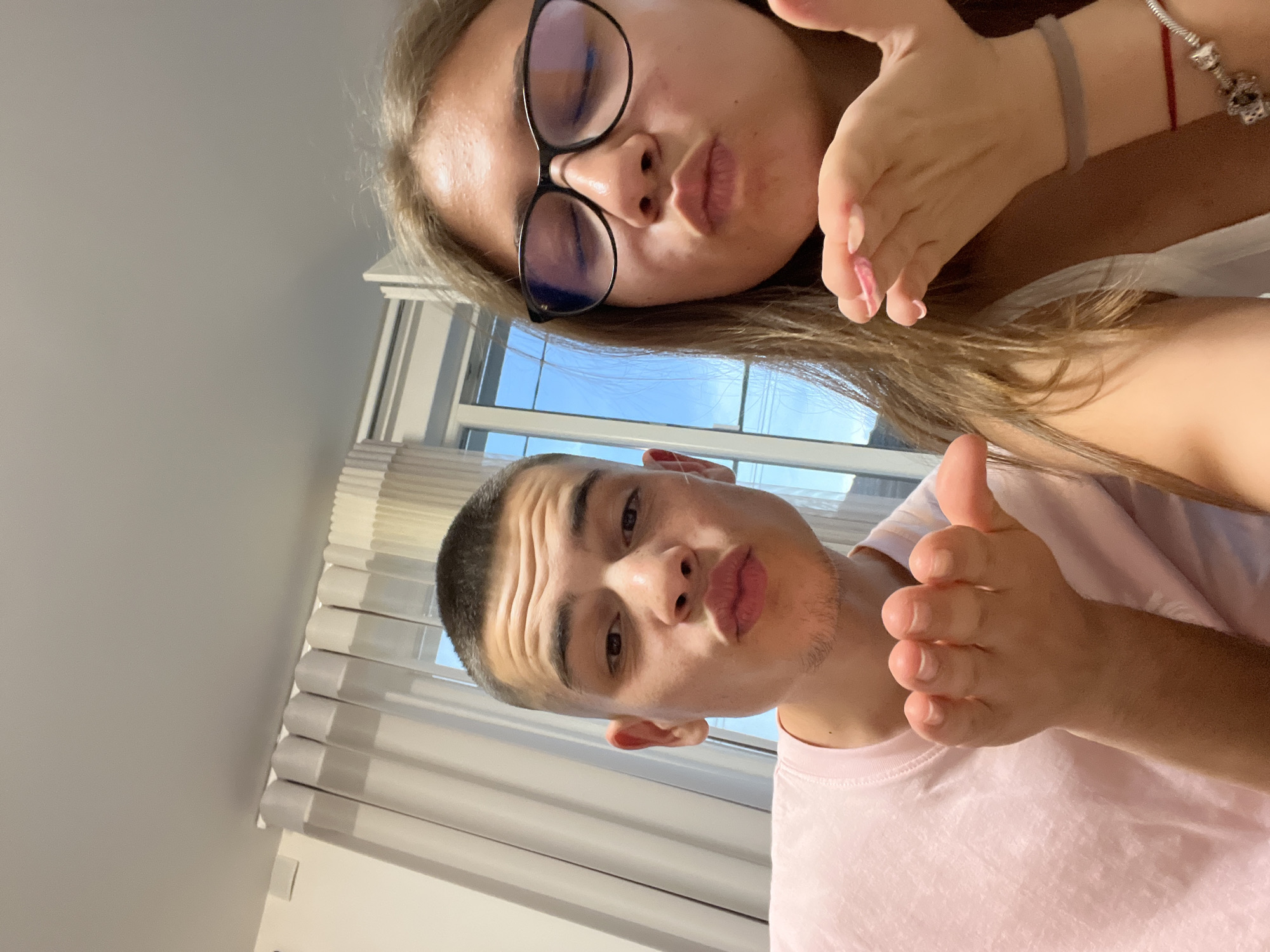 The image displays two young individuals, a male on the left and a female on the right, making identical "blowing a kiss" gestures directly towards the viewer in what appears to be a selfie. The scene is situated indoors, specifically in the city of Karavelovo, Bulgaria.
The male on the left has short, dark hair (buzz cut) and light stubble on his chin and jawline. He is wearing a light pink crew-neck t-shirt. His lips are pursed, and his right hand is cupped with fingers together, palm facing forward, in the kissing gesture. Horizontal wrinkles are visible on his forehead, consistent with his facial expression.
The female on the right has long, light brown hair, black-rimmed glasses, and appears to have some blemishes on her forehead. Her lips are also pursed, and her eyes are partially closed. She is wearing a white sleeveless top. On her left wrist, she has a grey elastic band, a thin red string bracelet, and a silver charm bracelet with multiple charms, including one dark element. Her left hand is cupped with fingers together, palm facing forward, mirroring the male's gesture, and her fingernails are painted with light pink polish.
The background shows a brightly lit room. To the left, a white wall features a small, white rectangular fixture mounted high near the ceiling, alongside vertically pleated, light-colored curtains. To the right, a multi-pane window is visible, revealing a clear blue sky, suggesting daylight. Similar light-colored curtains frame the window. The ceiling above is plain and light grey.