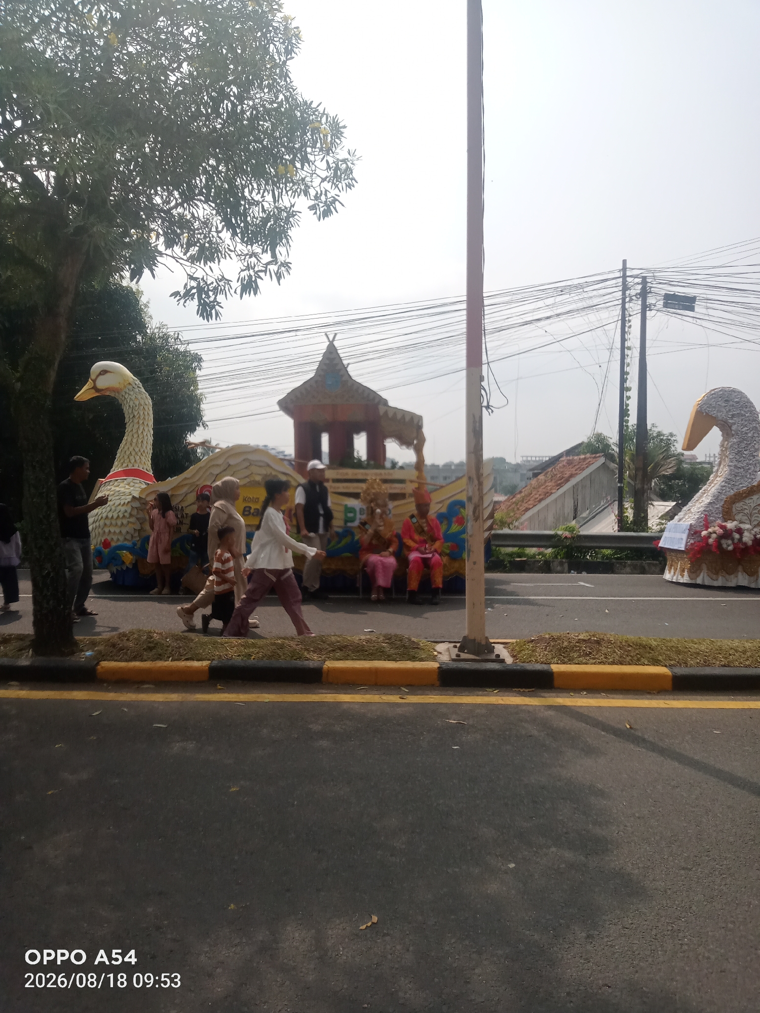 An outdoor daytime image shows a parade float on a paved road in Jambi, Indonesia. The float is decorated with two large swan figures. The swan on the left is facing left and appears to be made of a textured material with yellow accents. The swan on the right is facing right and is adorned with white fabric and red flowers. Between the swans is a smaller, ornate structure with a peaked roof, resembling a traditional Indonesian building, and a banner with text that is partially obscured. Several people are visible in and around the float. On the left, a man in a black shirt stands near the swan. To the right of the float, several people, including women in headscarves and children, are walking. Two individuals, dressed in colorful, traditional attire, are seated on the float. The environment includes a tree on the left, power lines overhead, and a slightly elevated walkway with grass and hedges in the background. The road is lined with a curb painted black and yellow. The image is timestamped "OPPO A54 2026/08/18 09:53".