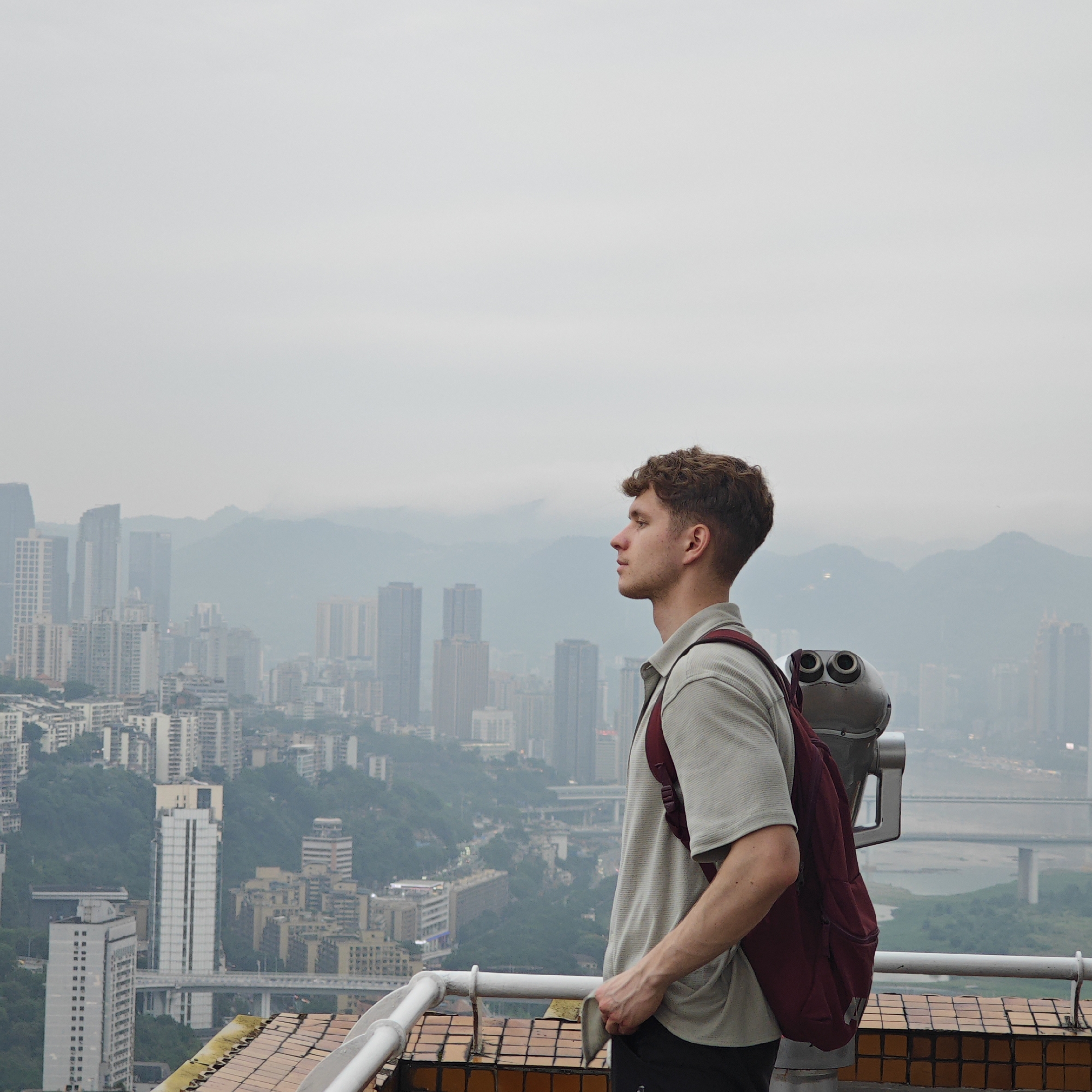 The photograph captures a man in a contemplative pose, looking out over a dense urban landscape. He stands on a rooftop observation point, evidenced by the presence of coin-operated binoculars and a metal railing. The man is fair-skinned with curly brown hair, wearing a collared shirt and a red backpack. He is positioned in the right foreground of the image, with his head turned to the left, gazing at the city skyline.
The backdrop is dominated by numerous skyscrapers of varying heights, densely packed together, suggesting a major urban center. In the distance, there are mountains shrouded in a light haze, indicative of some atmospheric pollution or fog. The sky is overcast, creating a soft, diffused light.
Based on the skyline and the presence of the river with several bridges, the location is identified as Yuzhong District, Chongqing, China. The observation point provides a panoramic view of the city's central business district and the surrounding geographical features.