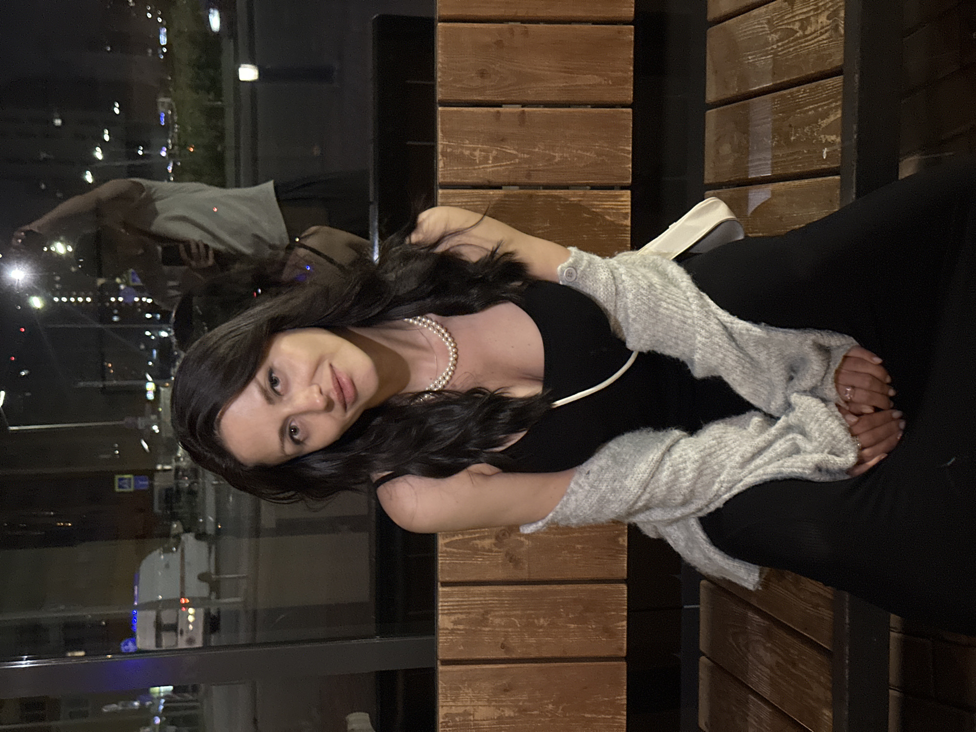 A medium-shot captures a woman seated on a wooden slatted bench at night, looking directly at the camera with a subtle smile. She has long, dark, wavy hair and is wearing a black sleeveless top/dress with thin straps, adorned with a double-strand pearl necklace. A light grey, possibly knitted cardigan, is draped over her shoulders and arms. Her hands are clasped in her lap, revealing manicured nails and rings. A white strap, likely from a bag, runs diagonally across her body from her left shoulder.
The scene is viewed through a glass panel, creating reflections. Prominently reflected is a male figure, presumably the photographer, wearing a light-colored t-shirt and dark bottoms, holding a smartphone up to take the picture. Beyond the reflection, a nighttime urban environment is visible. Streetlights, building lights, and a distant cityscape create a bokeh effect. On the left side of the background, a white ambulance-like vehicle with blue lights on its roof is parked or moving on a street, with another white car behind it. A yellow and blue street sign is also visible. The foreground seating area suggests an indoor or semi-enclosed public space, while the background depicts an active street scene in the city of Unknown, Unknown.