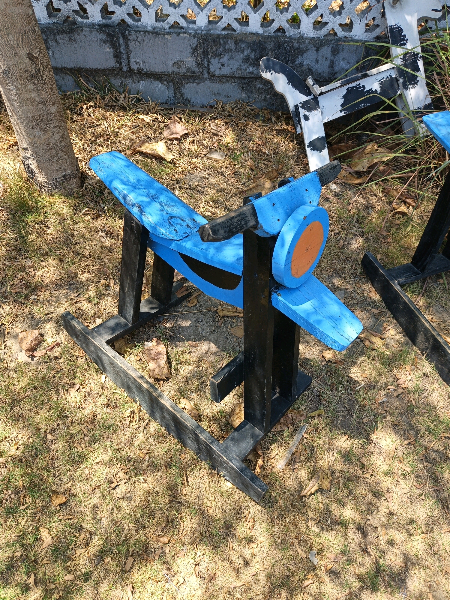 A blue wooden rocking airplane is positioned on dry grass and fallen leaves in front of a concrete wall with a decorative white fence. The airplane has black wooden supports and a propeller with an orange disc. To the right of the airplane, a portion of a black wooden structure is visible, partially obscured by grass. Further to the right, a black and white painted wooden rocking cow is situated behind the airplane. A tree trunk is on the left side of the frame. The overall scene is outdoors, with sunlight casting shadows on the ground.
