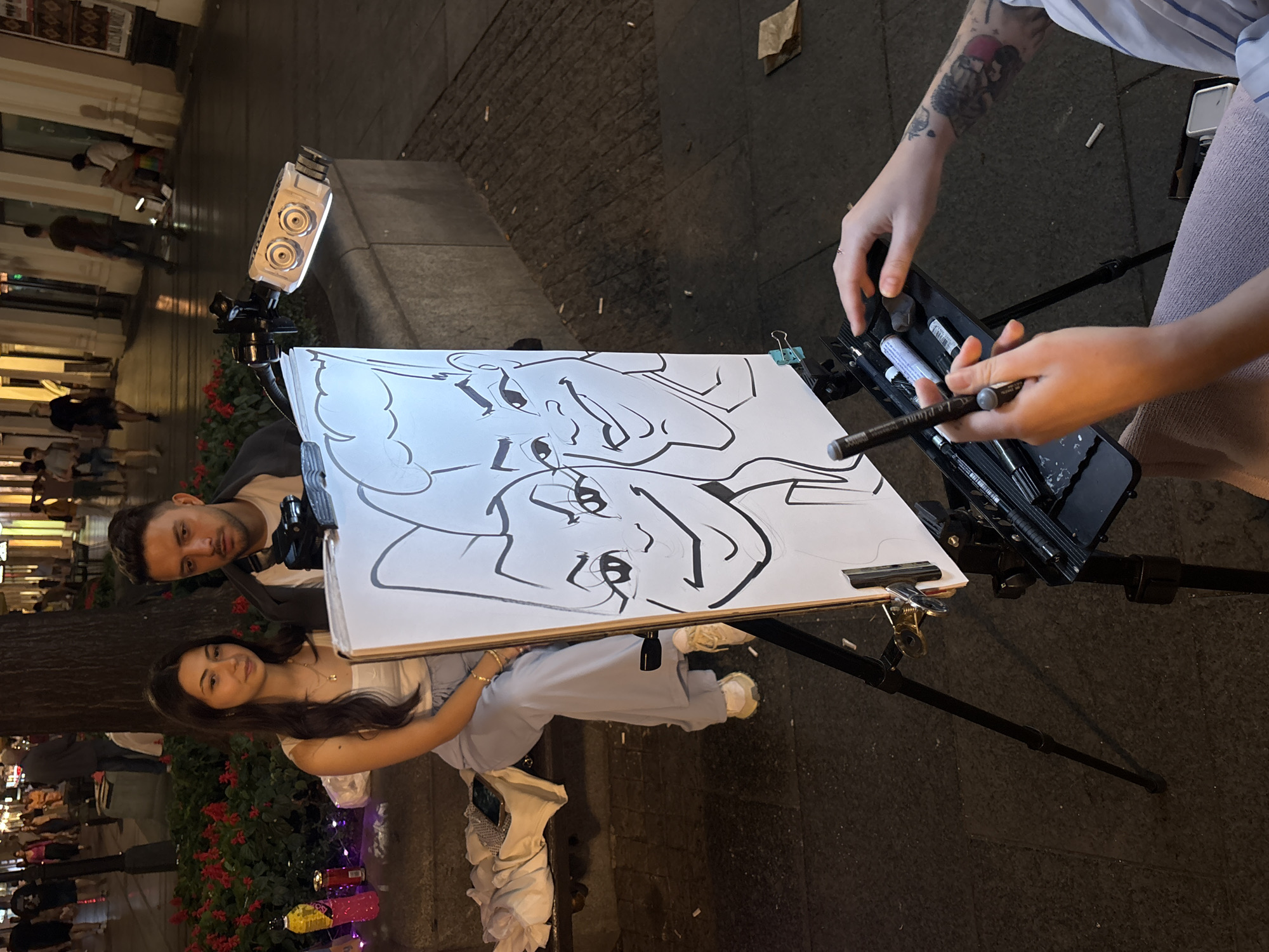 In Belgrade, Serbia, a young couple is seated on a bench, posed for a caricature artist who is drawing their likenesses. The woman, with long dark hair and a white tank top, sits to the left of the man, who wears a dark jacket over a light t-shirt. They are both looking slightly off-camera, while the artist, whose tattooed arm is visible in the foreground, is actively working on the drawing.
The drawing, displayed on an easel, features exaggerated, cartoonish depictions of the couple's faces. A portable light with two circular lenses is positioned on a ledge behind them, likely to illuminate the subjects. In front of the easel, a tray holds various art supplies, including pens and markers, accessible to the artist.
The scene takes place outdoors on a paved walkway, with a tree and red flowers providing a natural backdrop. In the background, blurry figures of pedestrians and shop fronts suggest a bustling urban environment, possibly a pedestrian street or promenade. The lighting indicates it is evening or nighttime.