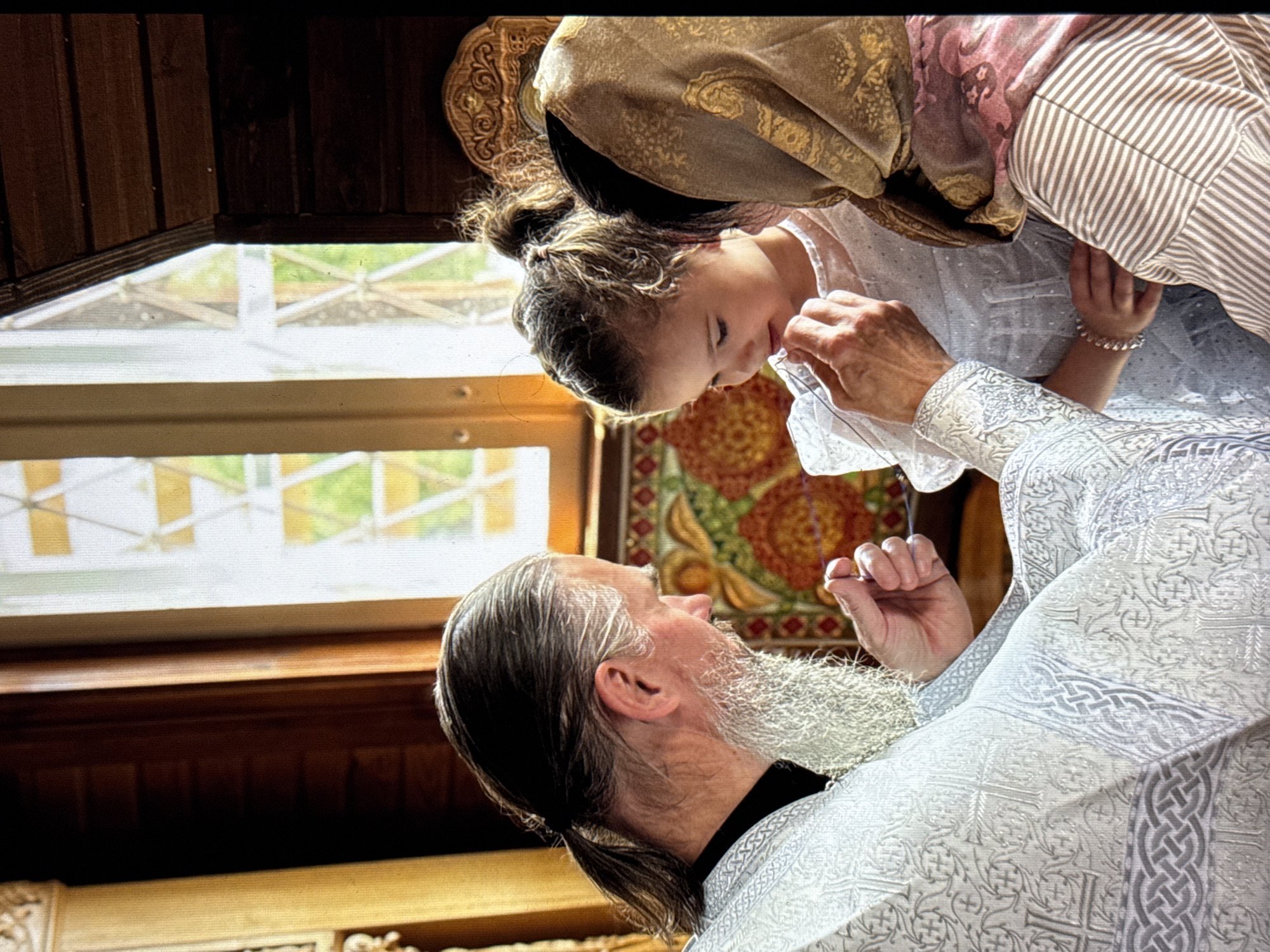 In an indoor setting, a bearded priest is holding a young girl, possibly during a religious ceremony. An older woman with a head covering stands behind the girl. The location is speculated to be in Minsk, Belarus, but there is no information in the image to support the location. A window is visible in the background, and there are decorated wooden elements around the window. The priest is wearing a white robe adorned with religious symbols. The girl wears a white dress and a bracelet. The older woman wears head coverings, possibly to denote a religious custom. The subjects appear to be in the middle of an interaction.