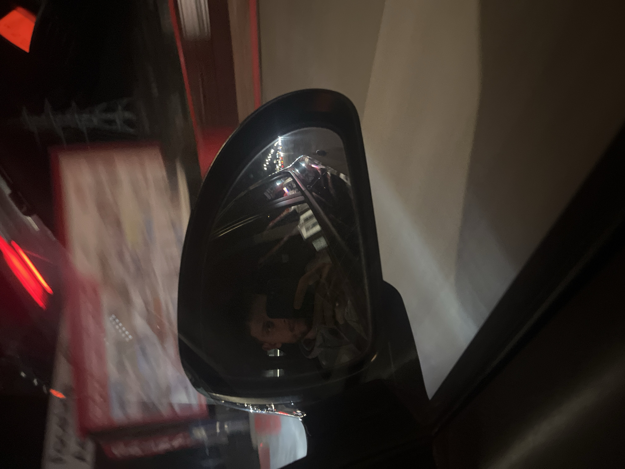 The image depicts a car's side mirror, reflecting a blurred urban street scene. The reflection includes building facades, signage, and indistinct figures. The mirror's housing is black, and the car's interior is visible in the background. The reflection suggests a nighttime environment with artificial lighting. The reflection appears to be in front of a store called "Masterhouse" in Sofia, Bulgaria. The reflection also captures the person taking the photo.