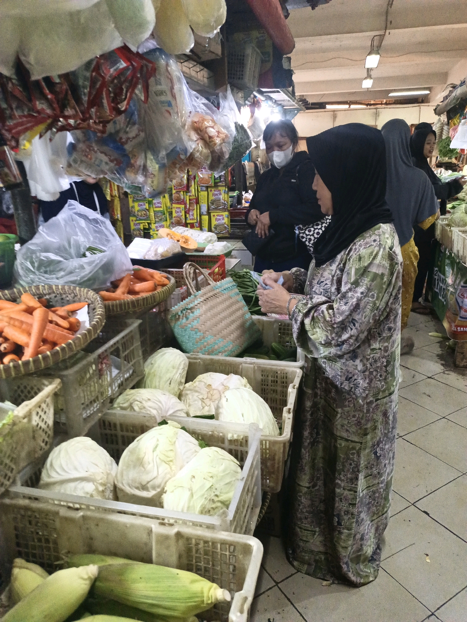 An outdoor market scene in South Jakarta, Indonesia, displays a variety of fresh produce and packaged goods. In the foreground, several white cabbages are arranged in plastic crates. To the left, a basket overflows with bright orange carrots. Further back, a woman wearing a black hijab and a patterned green and gray dress is examining money while standing near a crate of green beans. Behind her, another woman in a black hijab and mask appears to be interacting with her. Other market-goers are visible in the background, along with stalls filled with more produce and various bagged items hanging overhead. The ground is tiled, and fluorescent lights illuminate the busy marketplace.