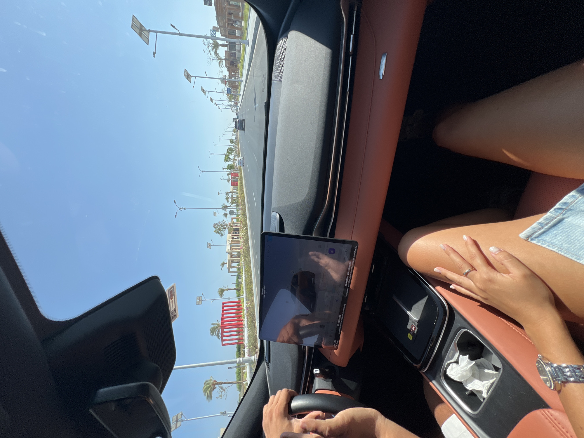 The image, taken from inside a car in Sharm El Sheikh, Egypt, captures a bright, sunny day. The perspective is from the passenger seat, looking forward through the windshield. The dashboard is visible, featuring a large central touchscreen displaying a navigation or car interface, with a reflection of a person's hand on it. The interior of the car appears to be luxurious, with tan leather seats and a dark dashboard.
Outside the car, a road stretches into the distance, lined with streetlights that have solar panels on top. Palm trees and modern-looking buildings with distinct red architectural features are visible along the roadside. The sky is a clear, vibrant blue. The road has a few cars driving ahead.
Two people are partially visible. One person's hands are on the steering wheel, suggesting they are driving. Another person's legs and a hand with a ring are visible on the passenger side, indicating two occupants in the front of the vehicle. The overall scene depicts a modern urban or resort setting under clear, sunny weather.